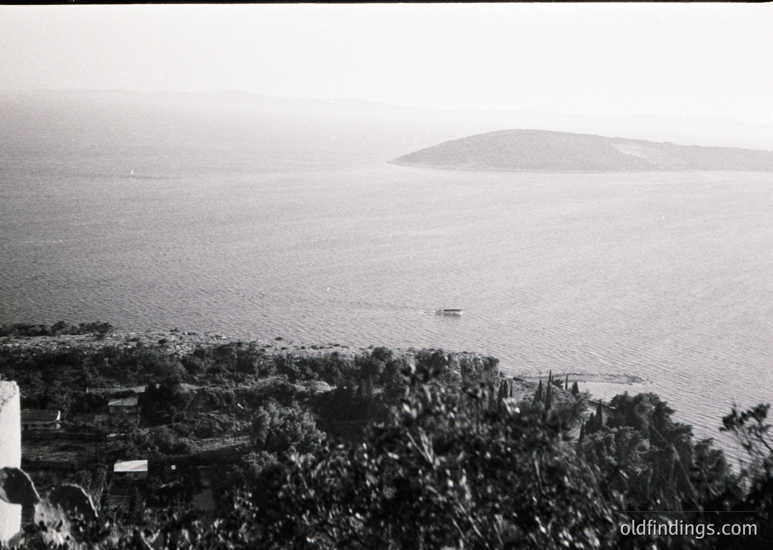 Black-and-white coastal scene featuring a lone boat on calm waters, framed by dense vegetation and low-lying structures. Distant island or landmass visible on horizon. Likely mid-20th century due to monochrome and composition style.