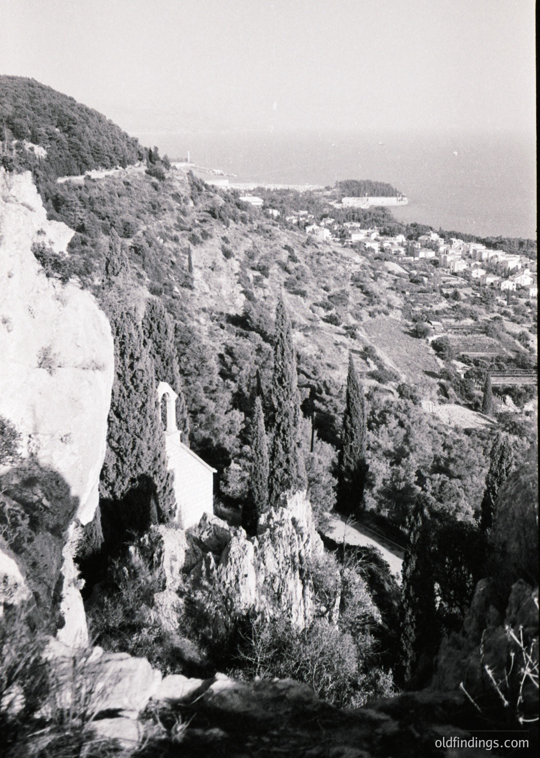 Black-and-white coastal landscape featuring rugged cliffs and a densely built seaside town. The rocky terrain dominates the foreground, leading to a densely packed urban area along the shore. The horizon reveals a calm sea and distant landmass. Likely mid-20th century based on architectural style and monochrome quality.