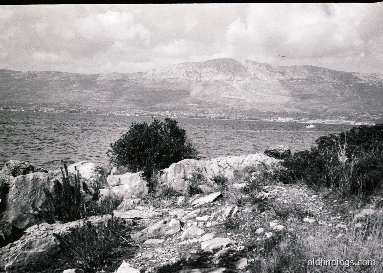 Black-and-white coastal landscape featuring rugged rocky foreground with sparse vegetation. Distant shoreline curves around a bay, with a faint outline of a coastal town or port. Mountain range in background, partially shrouded in mist. Likely Mediterranean or Black Sea region, mid-20th century.