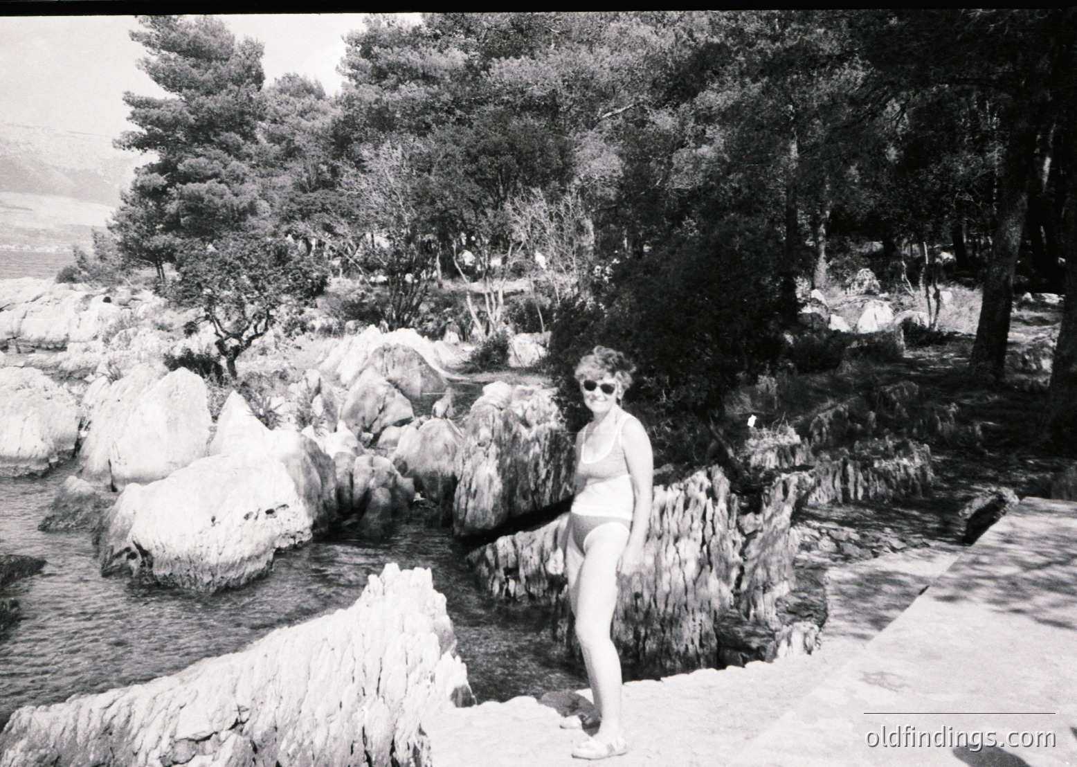 Black-and-white shot of a woman posing by a natural rock waterfall in a lush, forested setting. Mid-20th century attire (bikini, sunglasses, flat shoes) suggests a leisurely 1950s–1960s beach or resort scene. Geothermal or mineral-rich waters hint at a volcanic/thermal region.