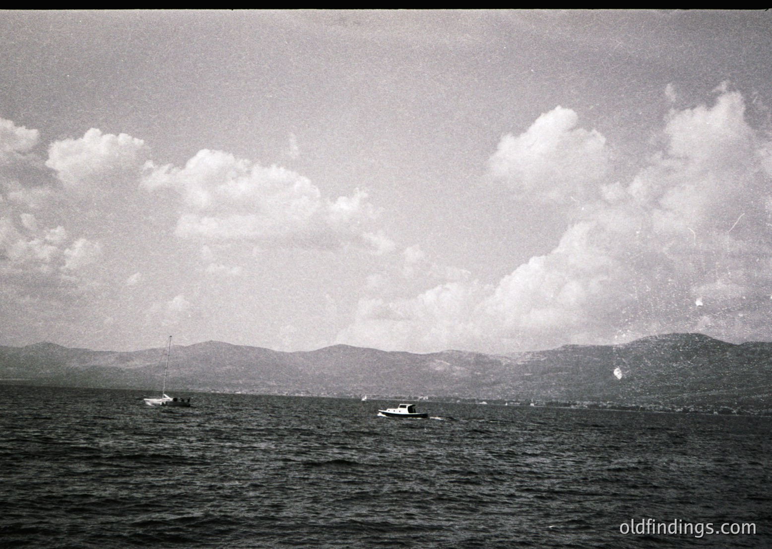 Vintage black-and-white seascape featuring two small boats on calm waters, with distant mountainous coastline under scattered clouds. Mid-20th century maritime or recreational scene, likely Mediterranean or coastal Europe.
