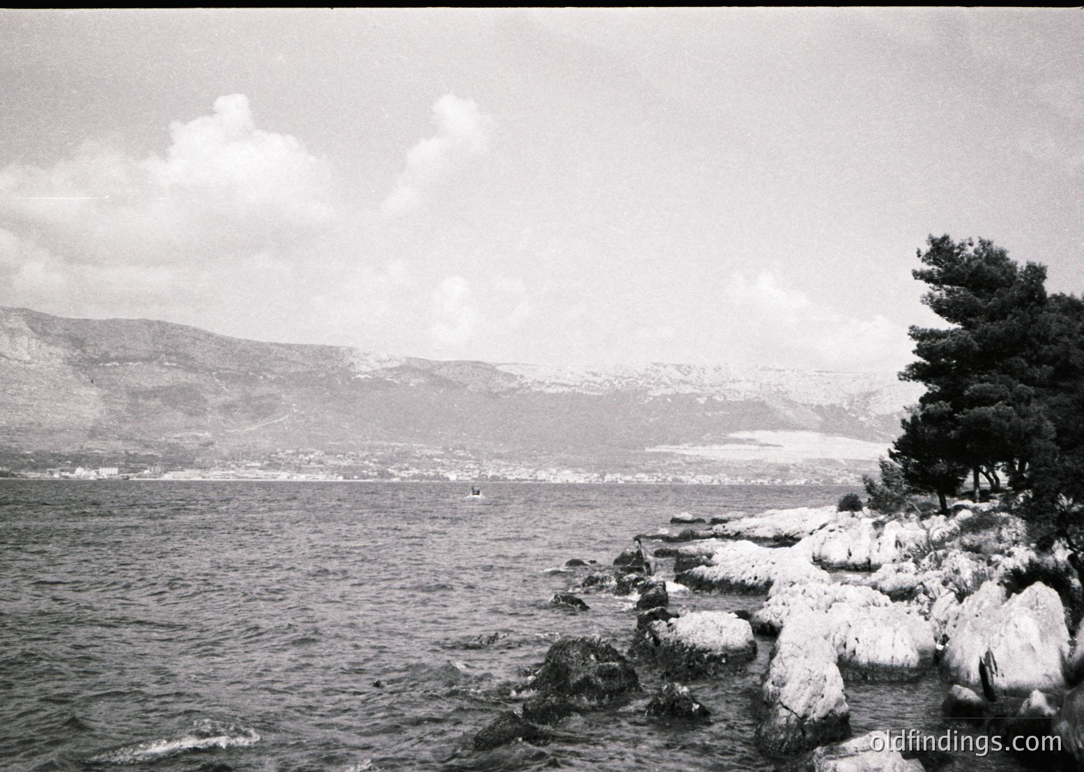 Mid-20th century black-and-white coastal scene: jagged rocks frame a rocky shoreline with minimal snow, while a lone boat drifts on calm waters. Distant hills and a coastal town stretch across the horizon under overcast skies.