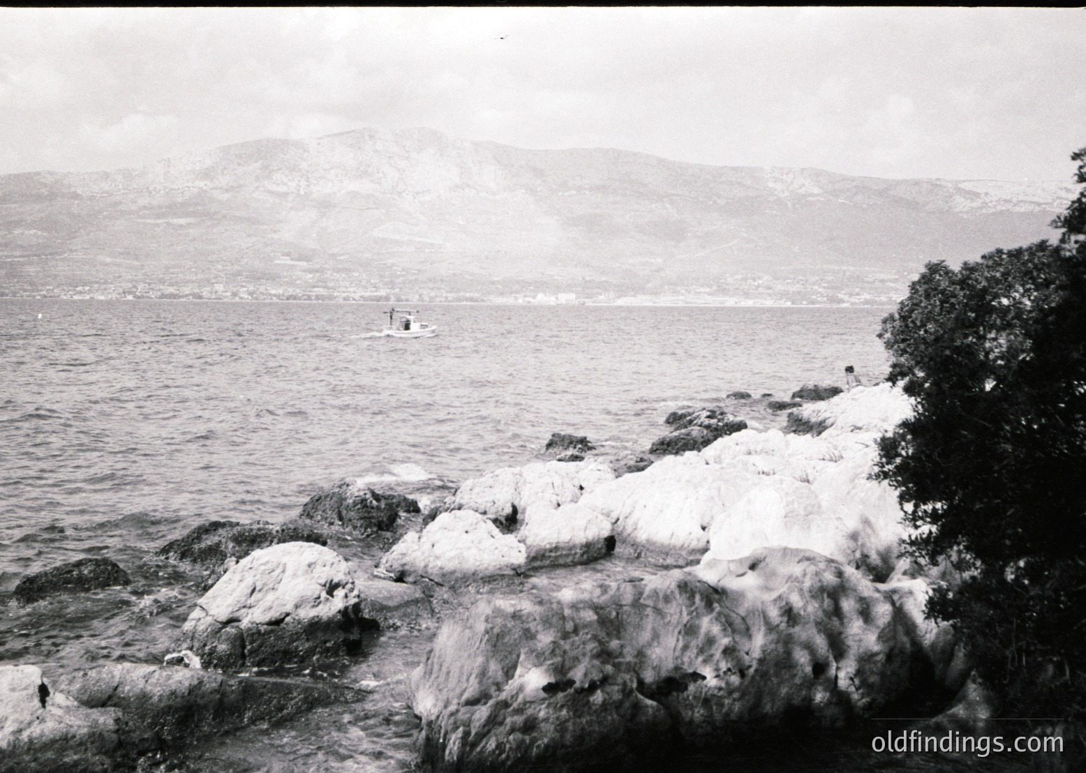Vintage black-and-white coastal scene featuring jagged rocks in foreground, a lone boat on choppy waters, and misty mountains in background. Likely early-to-mid 20th century maritime or seaside setting.