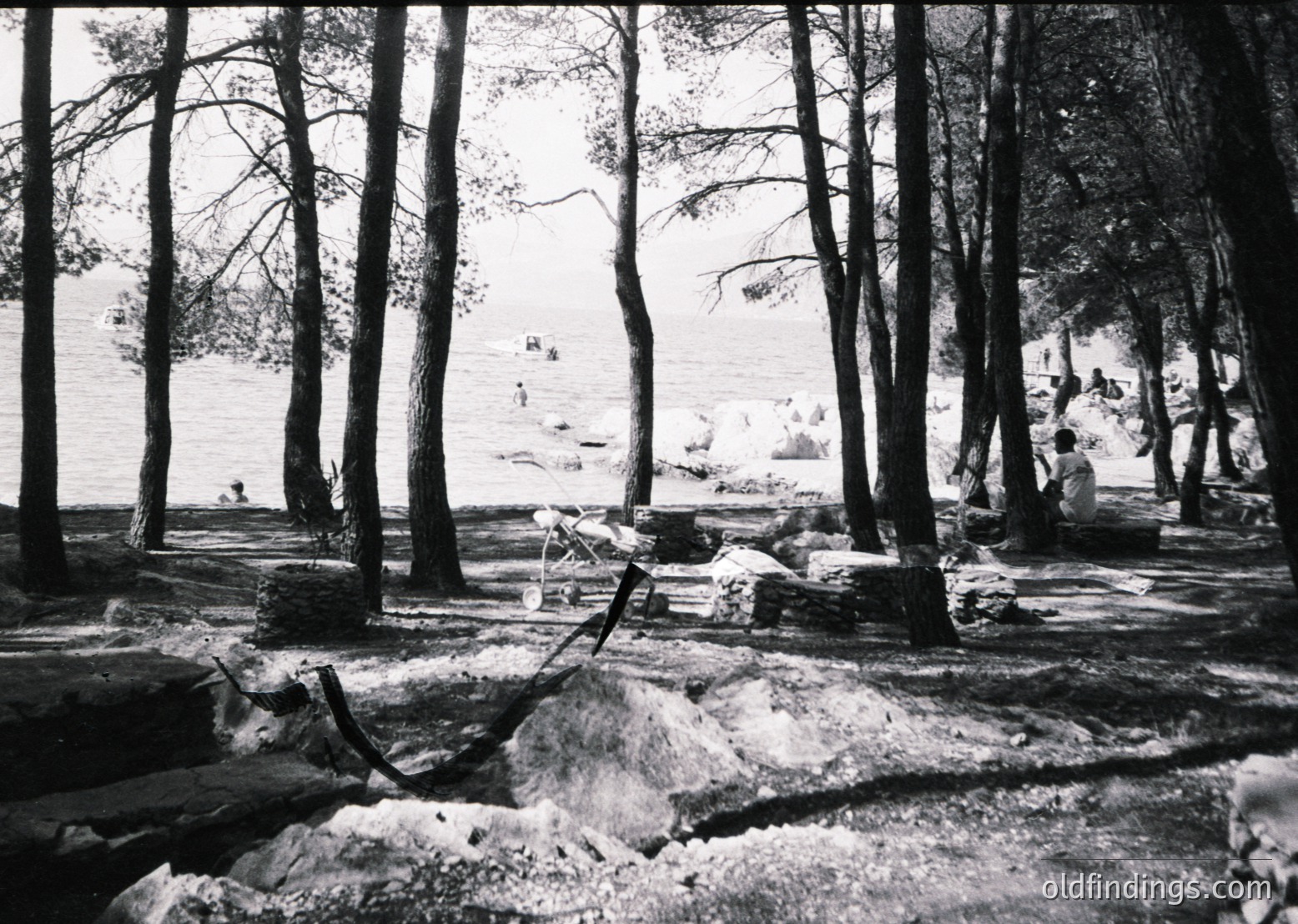 Black-and-white lakeside scene featuring dense forest framing a rocky shoreline. A lone figure sits on a rock near the water’s edge, while distant swimmers dot the lake. Mid-20th century vintage, likely , with natural lighting and minimal human intervention.