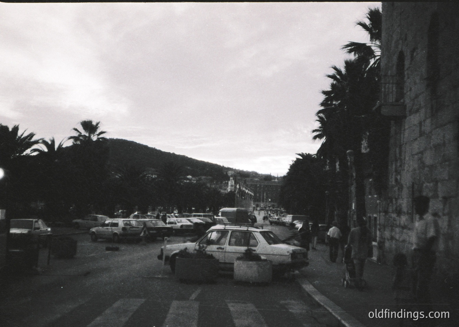 Mid-century coastal street scene with vintage cars parked along a promenade. Palm trees line the road, and a stone building occupies the right frame. Distant bridge and hilly terrain suggest a Mediterranean port town. Likely 1960s–1970s.