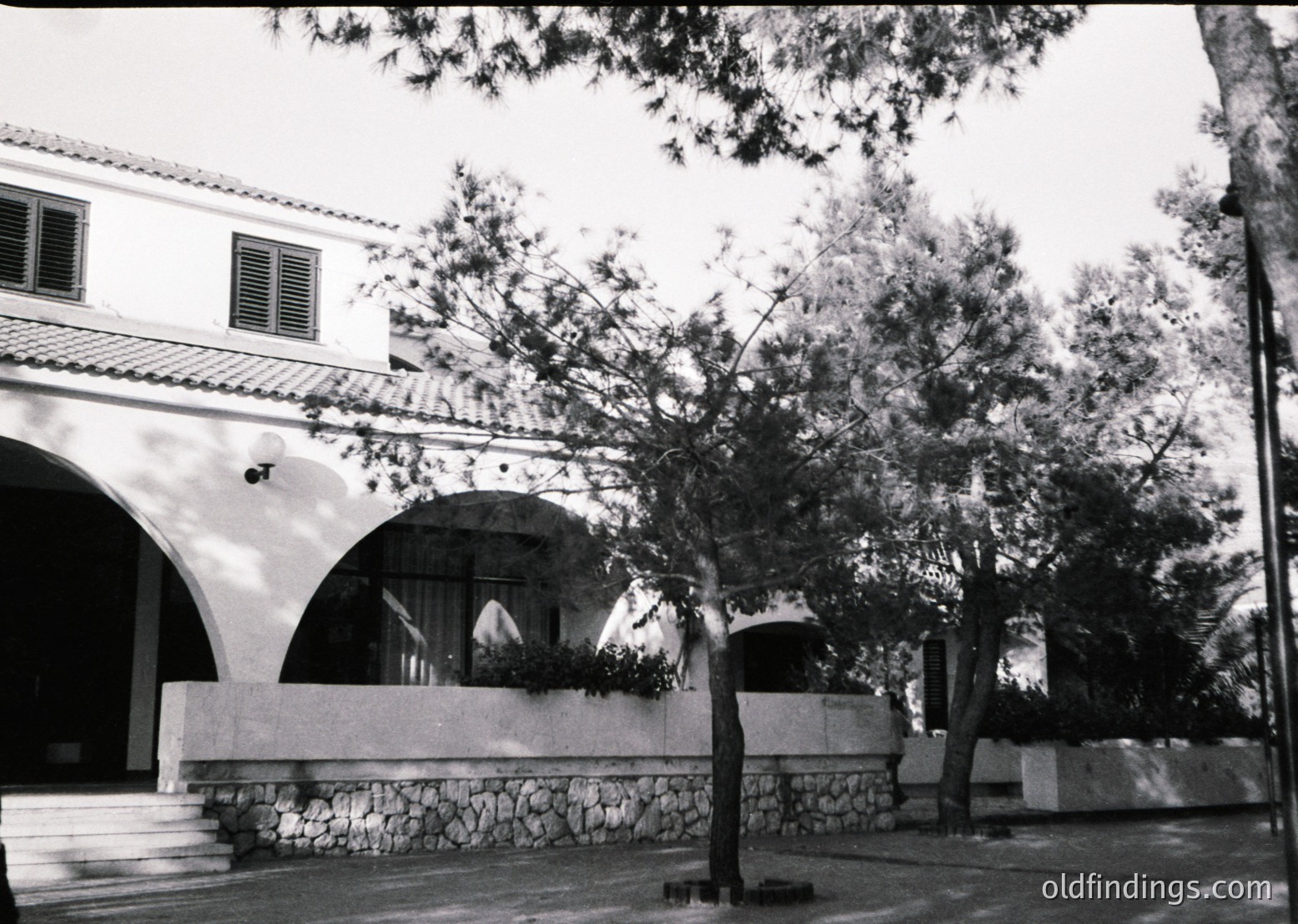 Mid-century Mediterranean-style courtyard with arched colonnade, stone base, and potted plants. Clean lines, minimalist design, and natural light suggest a resort or institutional building. Likely 1960s–1970s Mediterranean or coastal architecture.