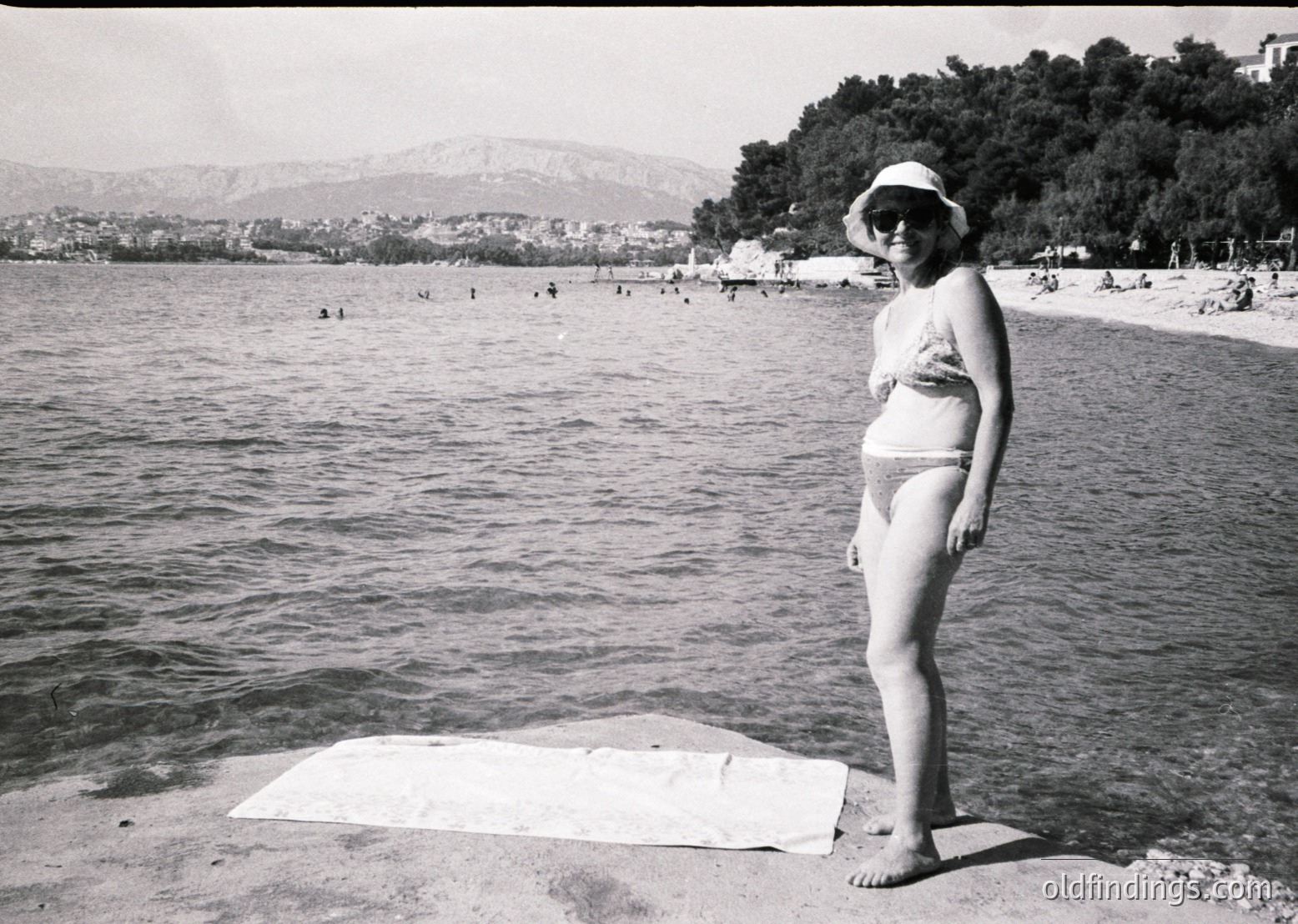 Black-and-white seaside portrait from mid-20th century, likely 1950s–1960s. Woman in a two-piece swimsuit poses on a towel near a crowded beach, with mountains and urban coastline in background. Classic retro beachwear and vintage photography style.