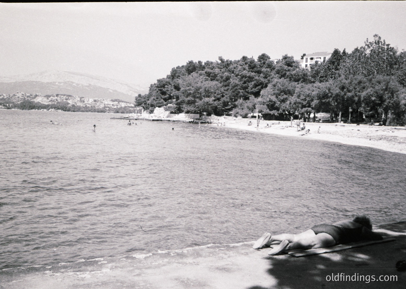 Black-and-white seaside scene featuring a crowded beach with scattered swimmers and sunbathers. Prominent rocky coastline with lush greenery and distant hills. Mid-20th century style beachgoers in swimwear. Likely Mediterranean coastal region.