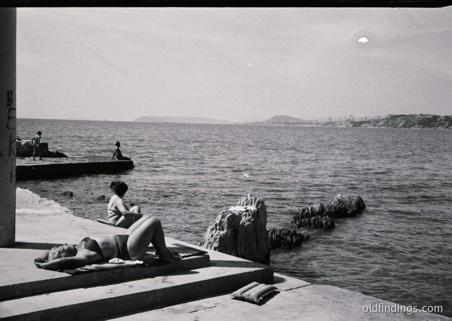 Mid-20th century seaside scene: two women sunbathe on concrete slabs beside a rocky breakwater. One reclines on a towel, the other sits upright. Distant city skyline and calm waters frame the composition. Likely Mediterranean or Black Sea coast, 1950s–1960s.