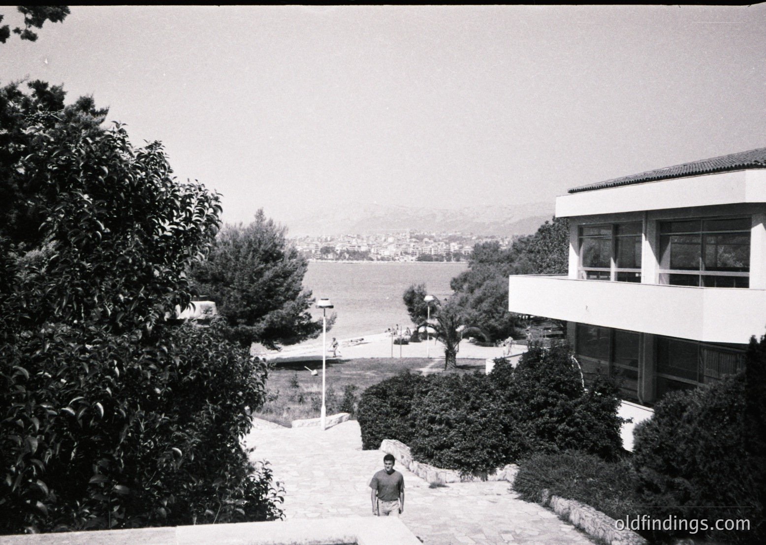 Mid-century seaside resort building with flat roof and large windows overlooking a calm coastal inlet. A man sits on stone steps near lush greenery, framed by mature trees. Distant hills and waterfront structures visible. Likely Mediterranean architecture.