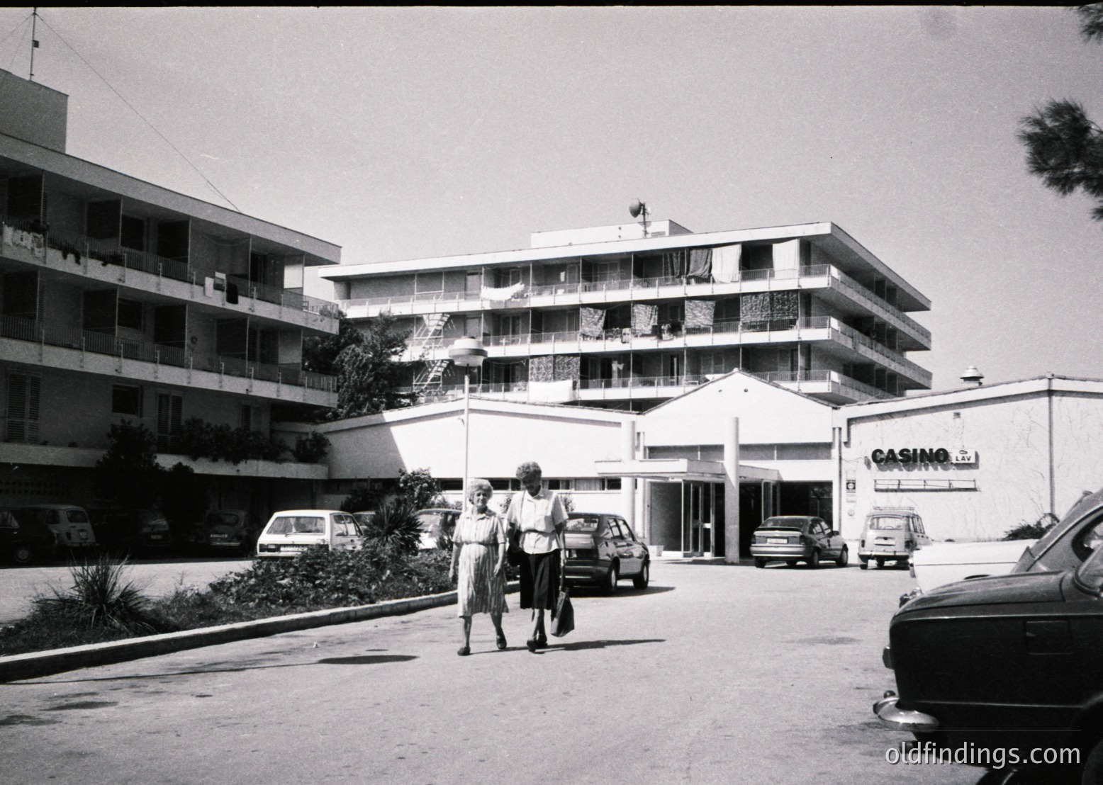 Mid-century concrete apartment block with flat balconies, flanked by a low-rise casino-style building (). Two pedestrians in light clothing cross a paved plaza near parked vintage cars (). Signage suggests a seaside resort atmosphere (). Overcast sky enhances the monochrome aesthetic ().