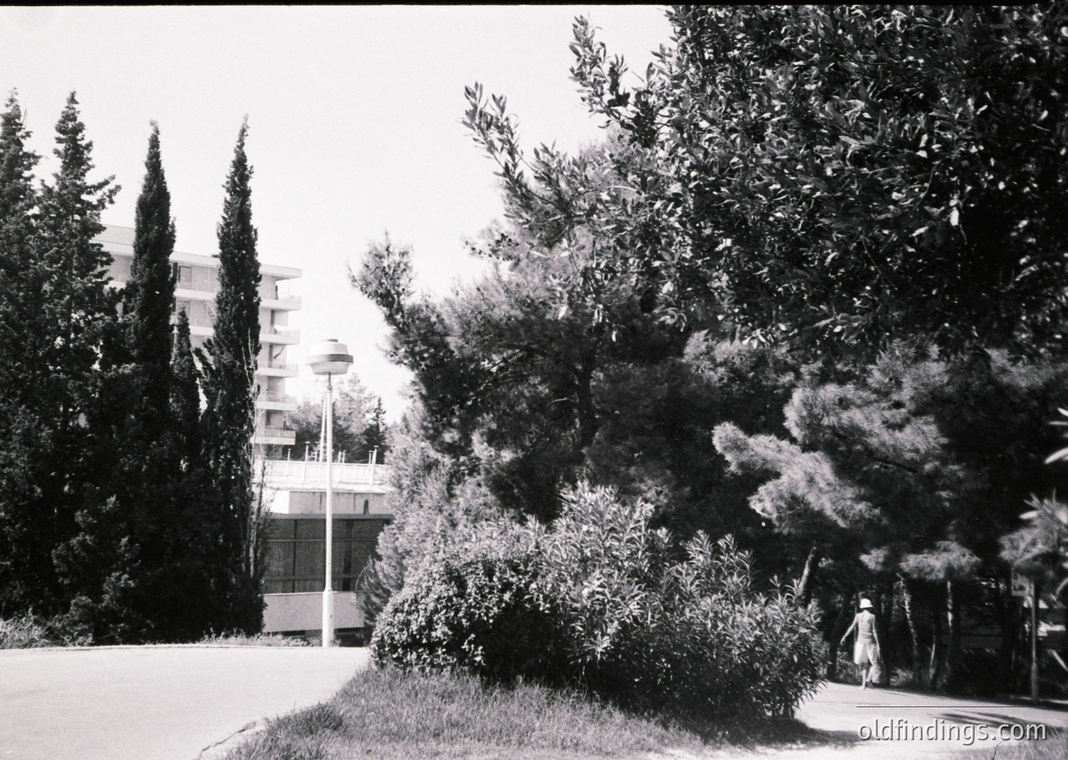 Mid-century modern seaside resort building with cylindrical balconies, framed by dense Mediterranean foliage. A lone figure in light clothing walks along a paved path, suggesting a relaxed, sunlit atmosphere. Likely Eastern European coastal architecture, 1960s–1970s.