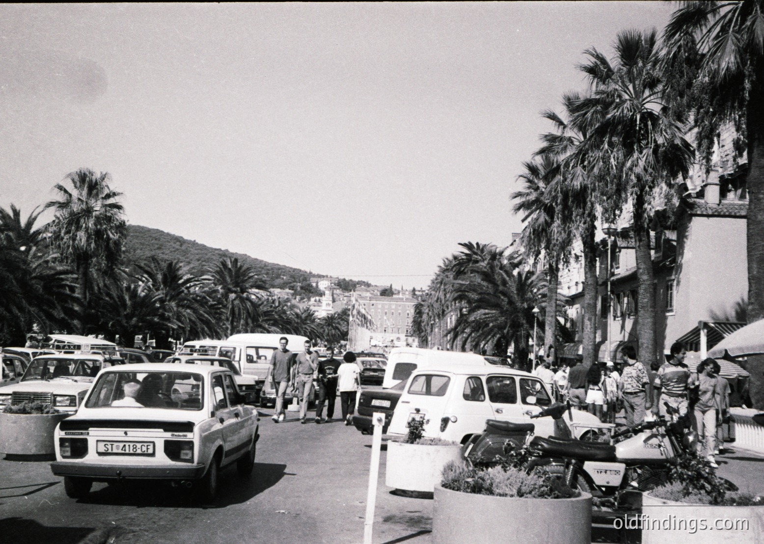 Mid-20th century seaside promenade with vintage cars, palm trees, and Mediterranean architecture. Crowded pedestrian area with parked vehicles and pedestrians in summer attire. Likely a European coastal town, possibly or , .