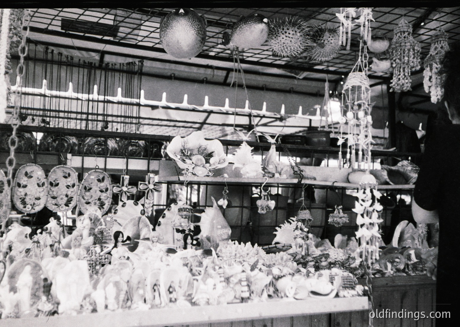 Vintage black-and-white display of handcrafted ceramic and glassware, likely from a 20th-century artisan fair or market stall. Shelves and hanging racks showcase intricately designed figurines, ornaments, and decorative items, including a large spherical ornament and a bust-like ceramic piece. The setting suggests a craft fair or exhibition hall, emphasizing artisanal production.