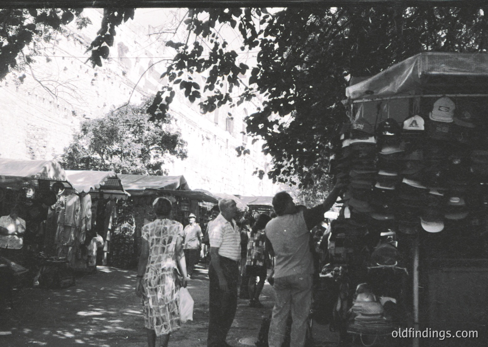 Vintage black-and-white outdoor market scene with stacked sandals and shoes for sale. Crowded stalls under leafy trees, featuring casual attire from mid-20th century. Likely Eastern European or Mediterranean setting.