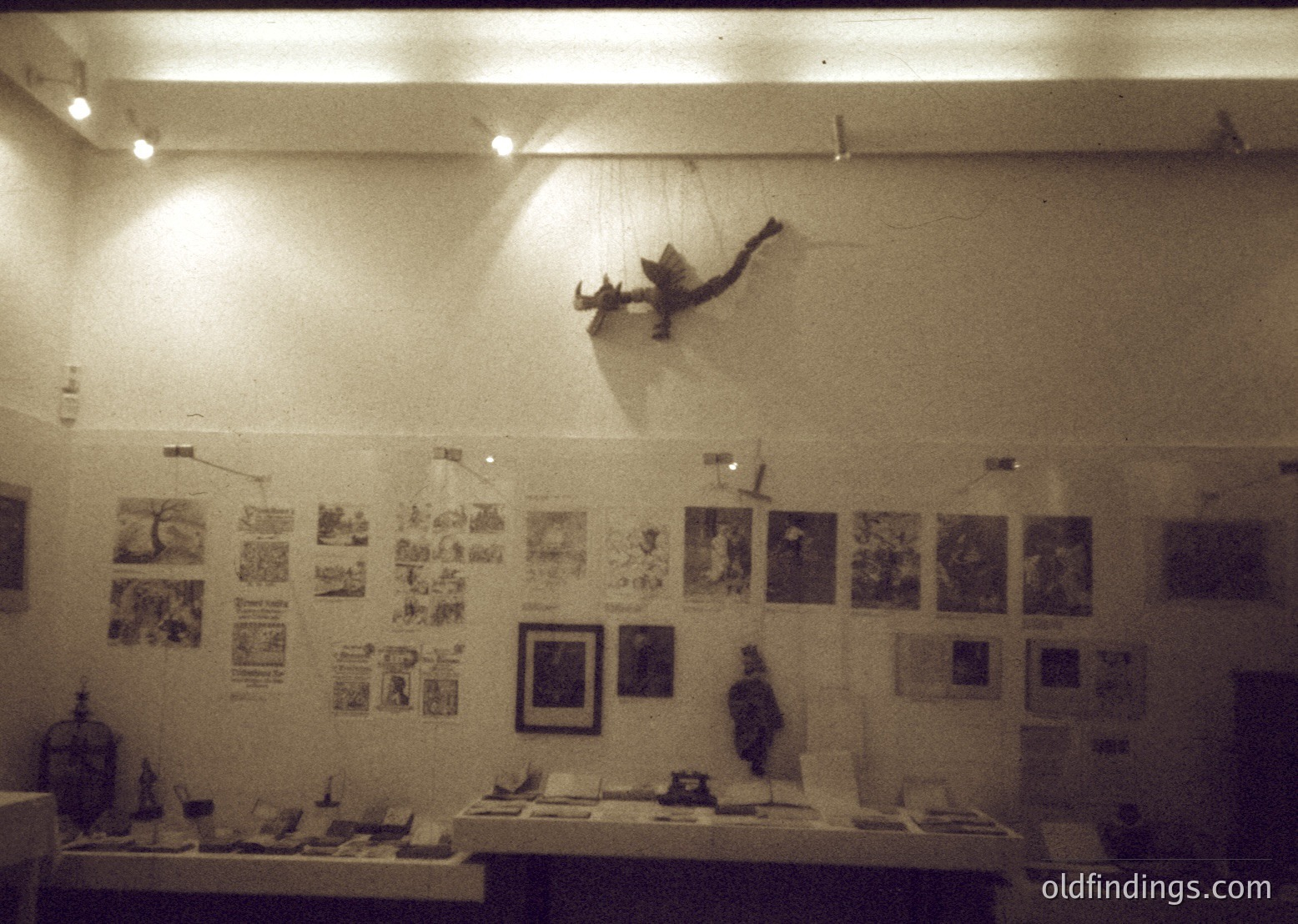 Vintage exhibition hall featuring framed posters, sculptures, and a suspended bird sculpture. Artwork includes black-and-white prints, political cartoons, and abstract designs. Display tables hold ceramic pieces and small figurines. Warm, ambient lighting enhances the nostalgic atmosphere. Likely a mid-20th century cultural or historical exhibit.