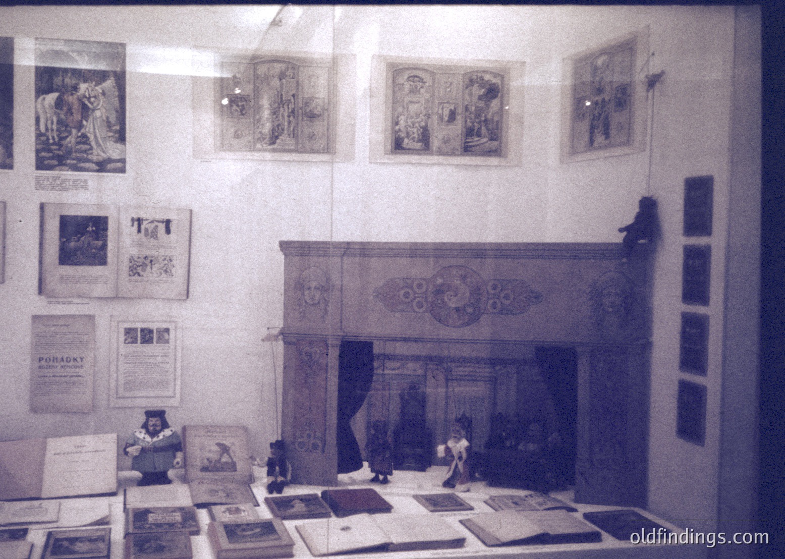 Vintage interior display featuring ornate fireplace with decorative surround, framed vintage posters, and miniature figurative scenes. Stacked books and documents on a table below. Likely a museum or historical exhibit from the mid-20th century.