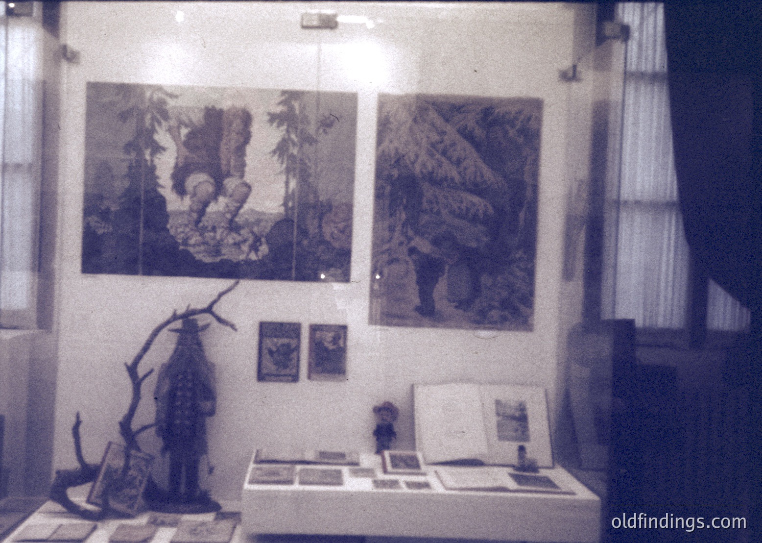 Vintage exhibition display featuring two large monochrome prints of forest scenes with human figures, likely from the 1960s–70s. Below, a table showcases smaller framed artworks, a taxidermied bird, and a wooden branch sculpture. Lighting highlights the curated arrangement.