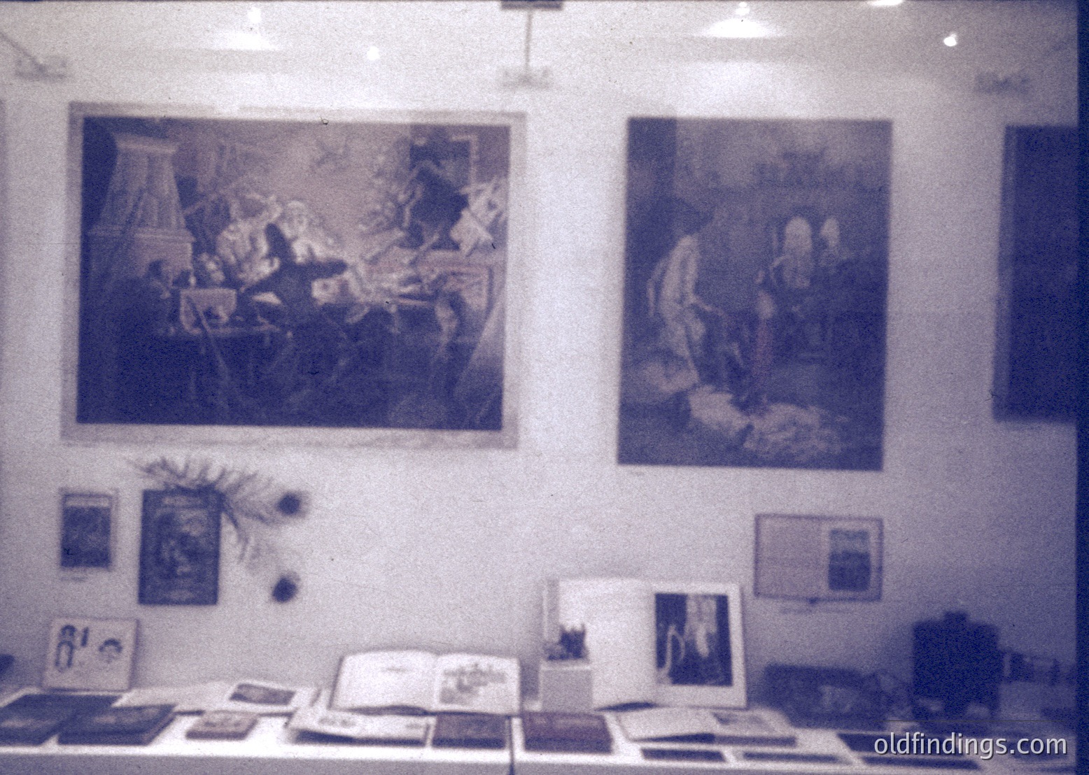 Vintage exhibition space featuring two sepia-toned classical paintings depicting historical/religious scenes. Below, framed artworks, peacock feather decor, and open books on a wooden table. Minimalist white walls and soft lighting evoke 1960s–1970s gallery aesthetics.