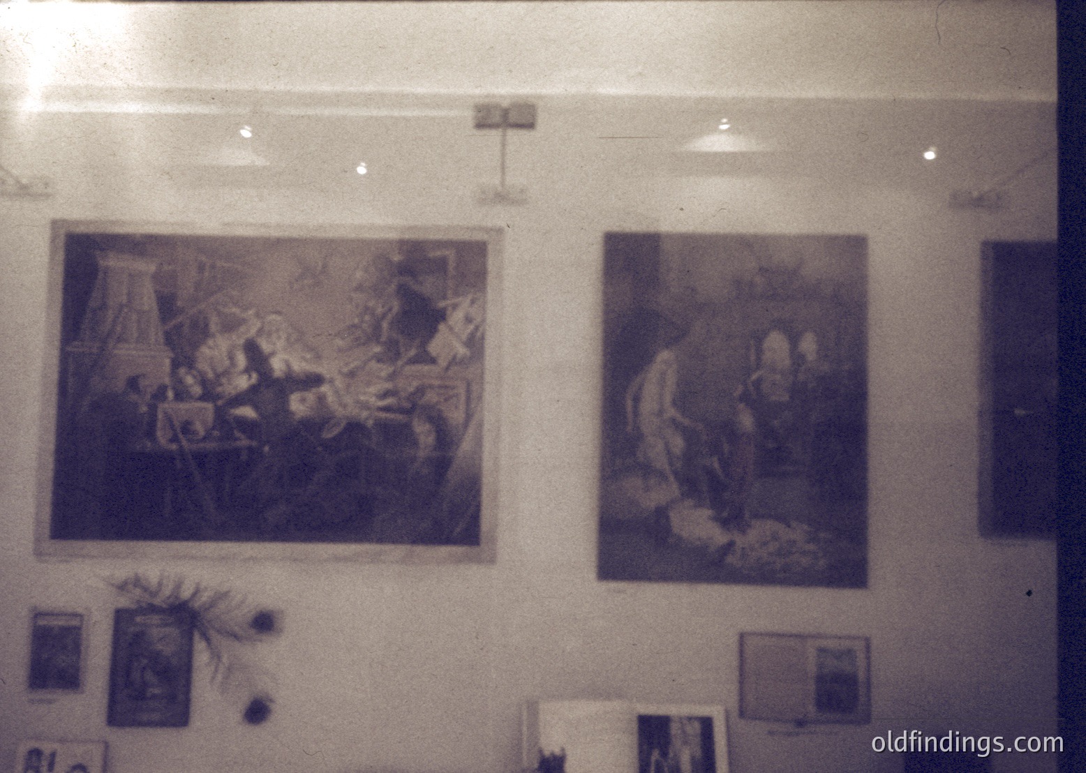 Vintage-style wall featuring two framed classical paintings depicting historical/religious scenes. Left: chaotic battle or siege with soldiers, horses, and crumbling architecture. Right: seated figure in flowing robes, possibly a biblical or mythological figure. Minimalist decor with a single potted plant and framed documents below. Likely 1970s–1990s institutional or public space.