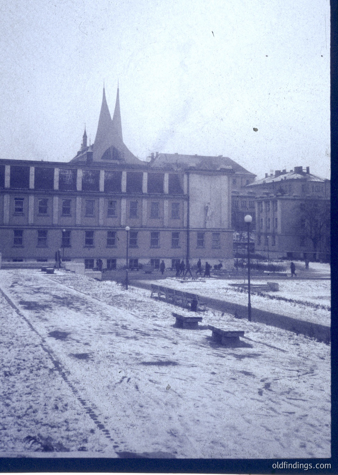 Snow-covered urban plaza with grand neoclassical building featuring twin spires and symmetrical façade. Pedestrians in winter attire, lampposts, and paved pathways. Likely Eastern European city, mid-20th century.