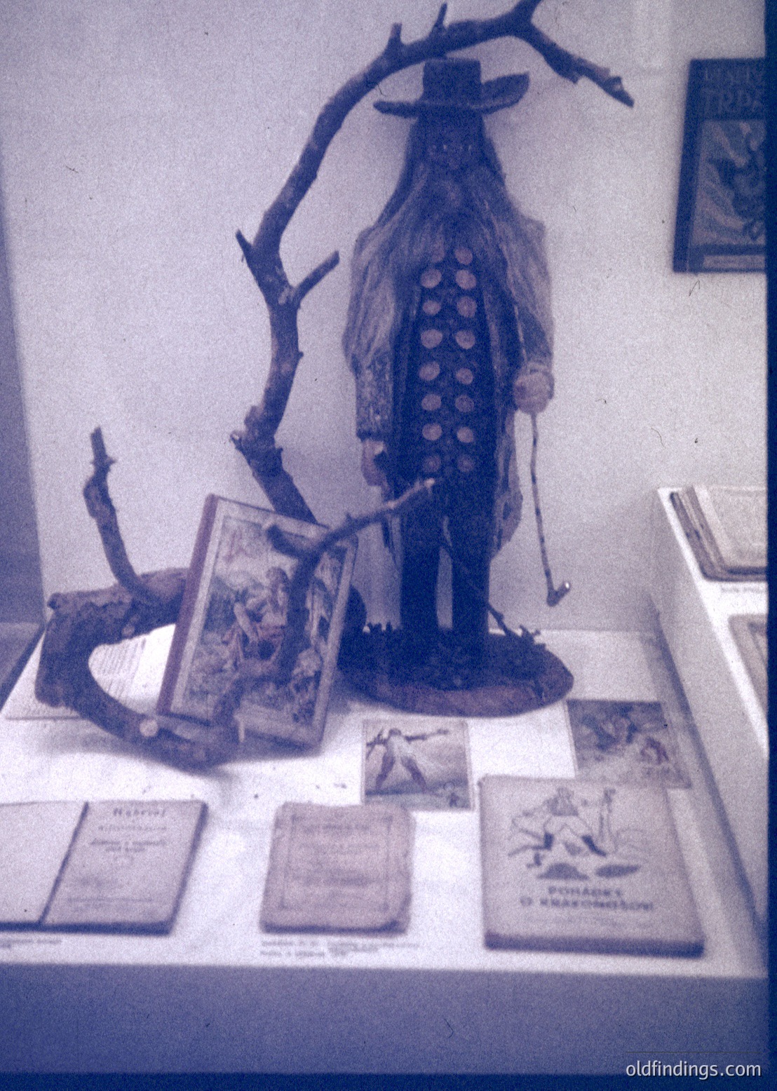 Vintage wooden scarecrow display with vintage books and rustic props. The scarecrow wears a wide-brimmed hat and a patterned dress, likely from Eastern European folklore. Books feature Cyrillic script, suggesting a Slavic cultural context. Displayed in a museum or exhibition setting, likely from the 20th century.