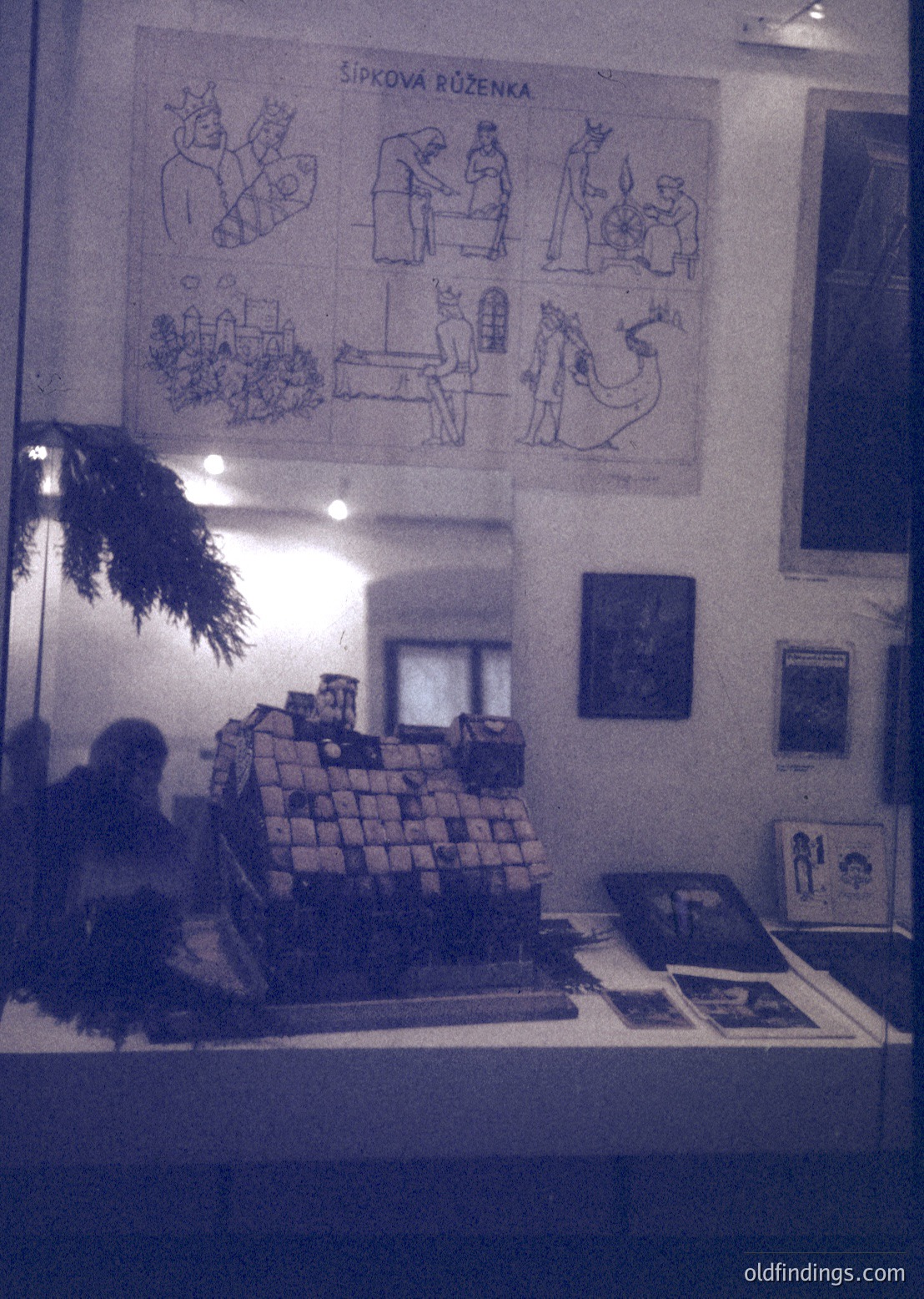 Vintage interior featuring a stacked brick display case with framed illustrations titled *"Šípková Růženka"* (Cinderella-inspired). Warm lighting highlights a blurred figure seated behind the counter, surrounded by framed art and holiday greenery. Likely a 1960s–1970s Czechoslovakian café or cultural exhibit. éDesign