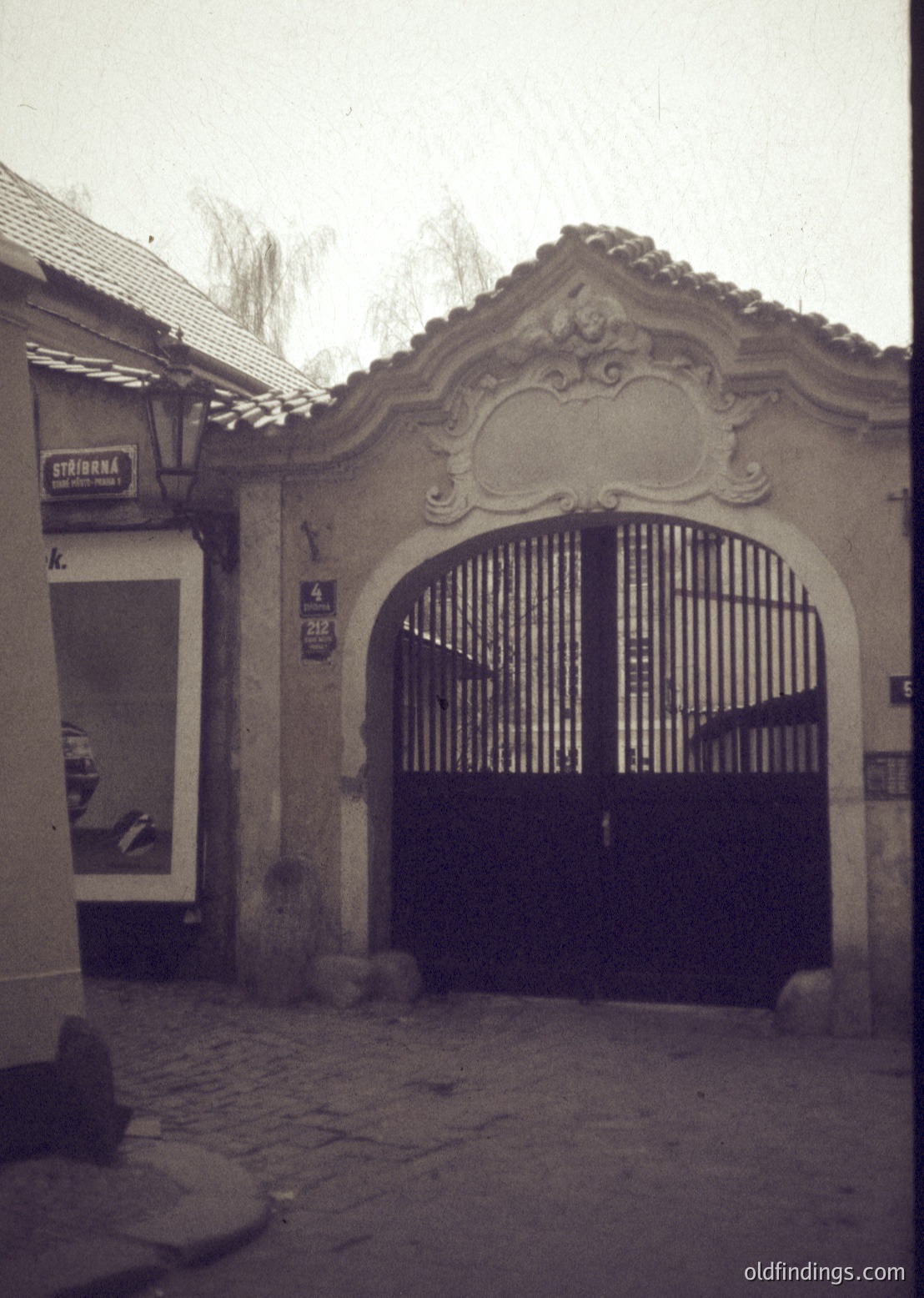 Vintage-style arched gate with ornate plasterwork and wrought-iron double doors, set in a cobblestone courtyard. Adjacent signage reads "STAIONA" (likely a shop or café). Architectural details suggest Eastern European influence, possibly Bulgaria. Black-and-white or sepia-toned, evoking mid-20th century charm.