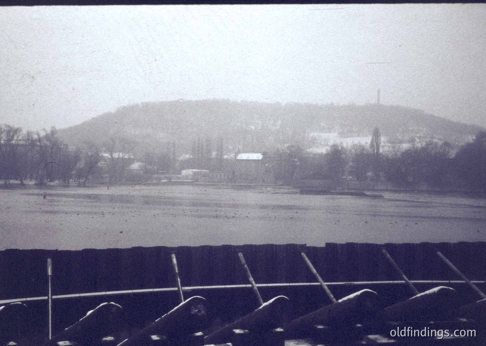 Vintage sepia-toned landscape featuring a rural scene with snow-covered fields and sparse trees. Distant hilltop with a lone structure, likely a radio tower. Foreground shows a fence with angled metal bars, suggesting a train or platform. *(Note: Exact location/time indeterminate due to lack of identifiable landmarks, but style suggests mid-20th century.)*