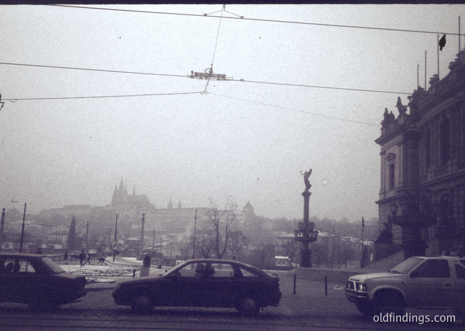 Vintage black-and-white street scene in a European city, likely Prague, showcasing historic architecture with a prominent column monument in foreground. Tram wires and early-model cars suggest mid-20th century (1950s–1960s). Foggy skyline reveals Gothic-style buildings, including the iconic Prague Castle.