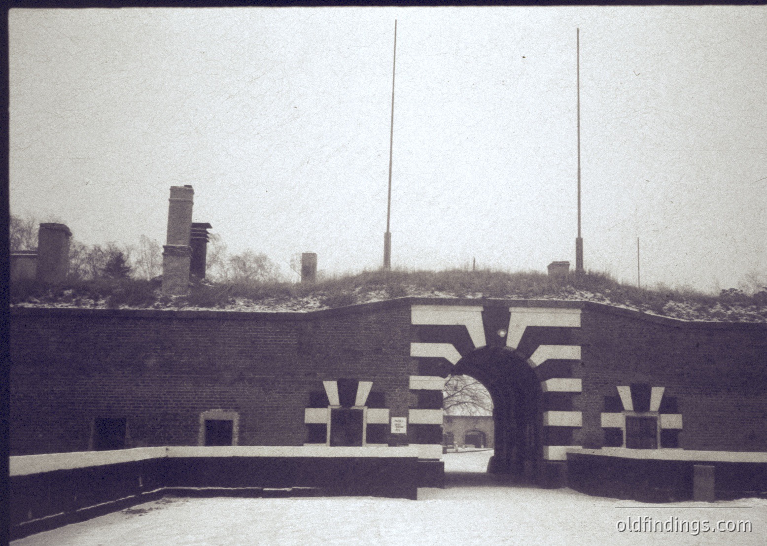Historic brick fortification entrance with "V" symbol, likely WWII-era German military architecture. Snow-covered ground and camouflaged rooftops suggest winter conditions. Industrial chimneys and antennae in background.