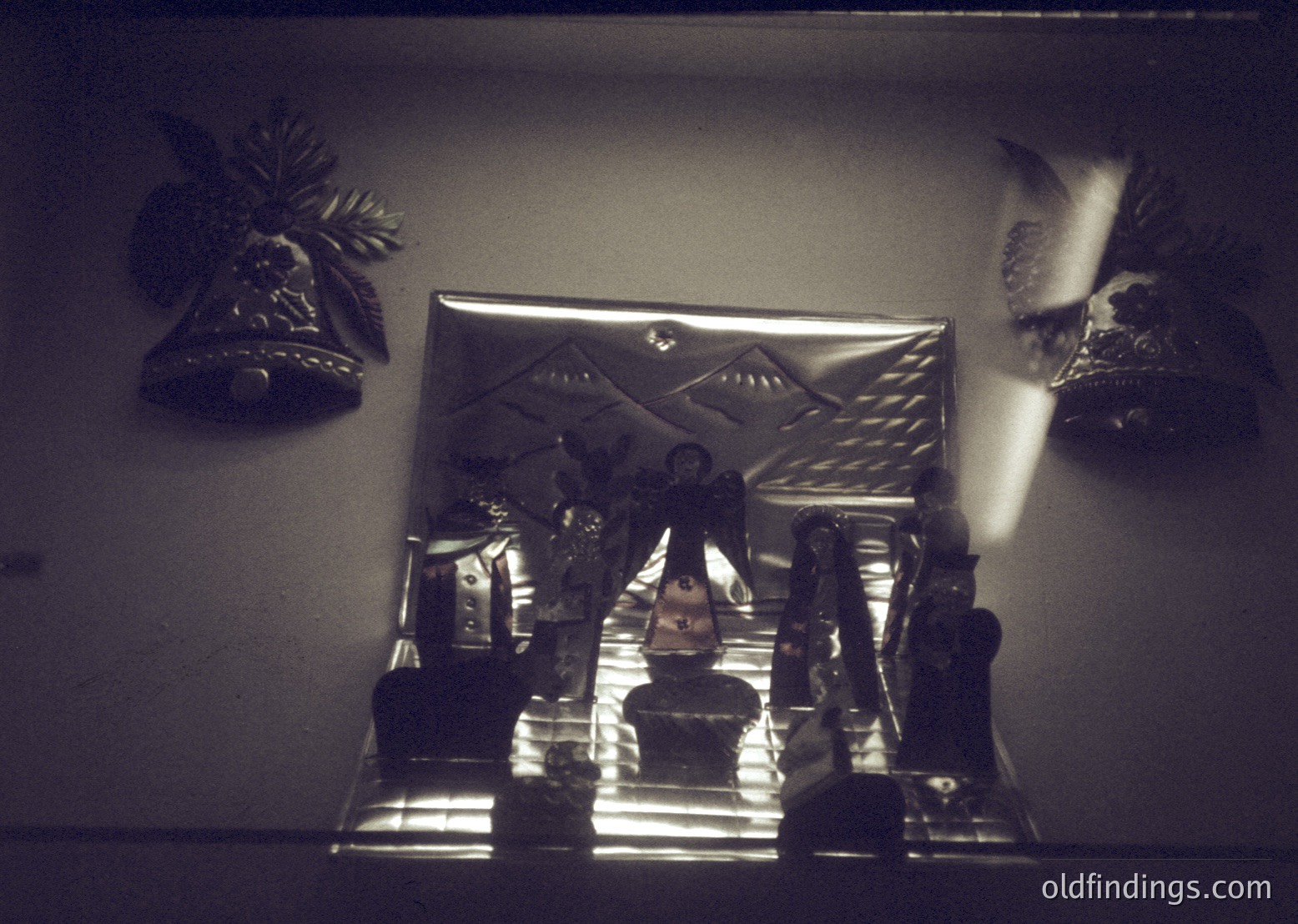 Vintage framed mirror with ornate metal frame featuring geometric patterns. Decorative wall bells and bird motifs flank the reflection of a seated figure in a dimly lit interior, suggesting a 1970s-1980s Eastern European home. The composition highlights mid-century interior design and folk-inspired decor.