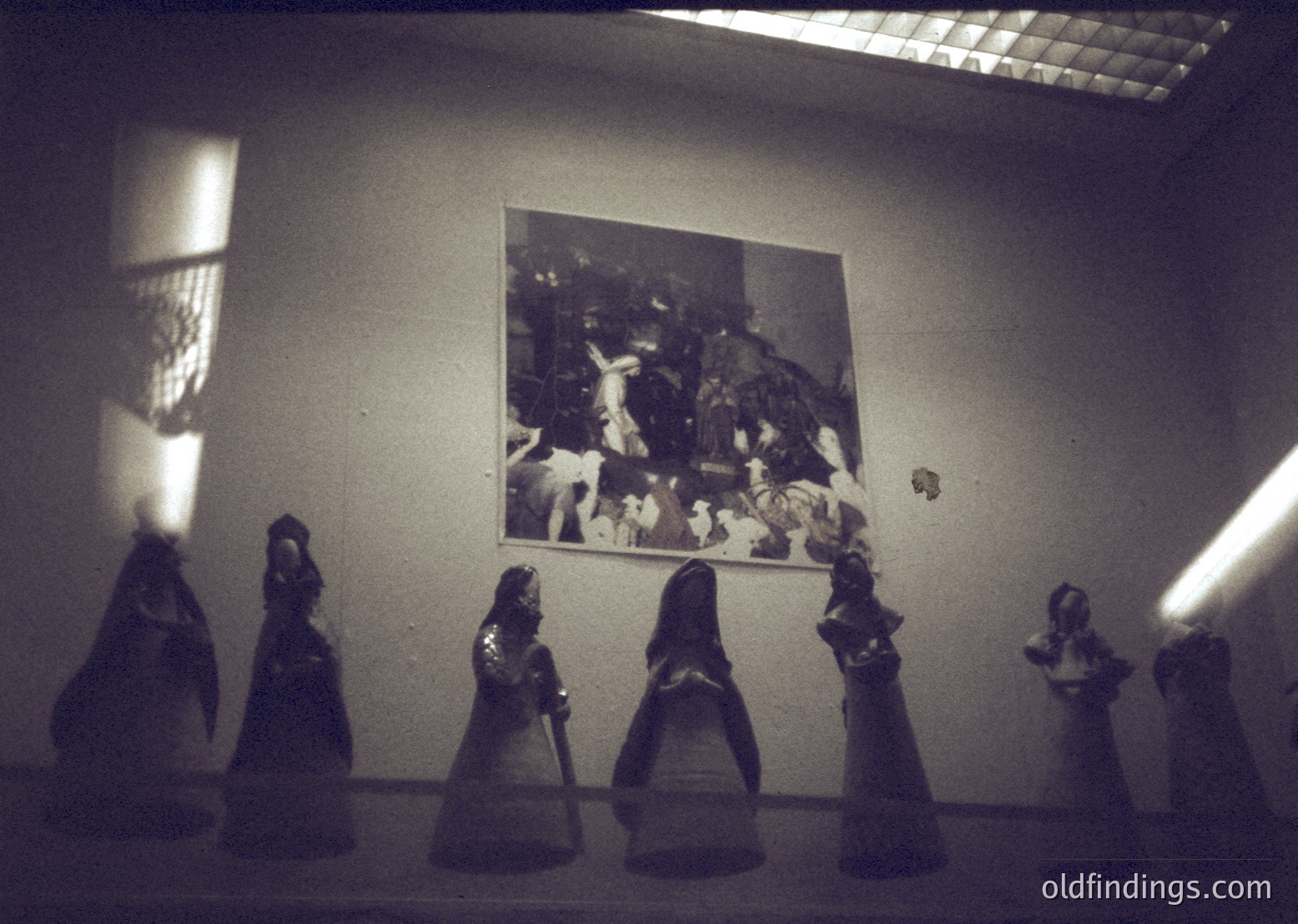 Vintage sepia-toned photo of a chess game in progress, with six chess pieces (likely pawns) in sharp focus. A framed black-and-white image of a crowded, lively party or gathering hangs on the wall behind. Warm light casts dramatic shadows, suggesting an intimate indoor setting.