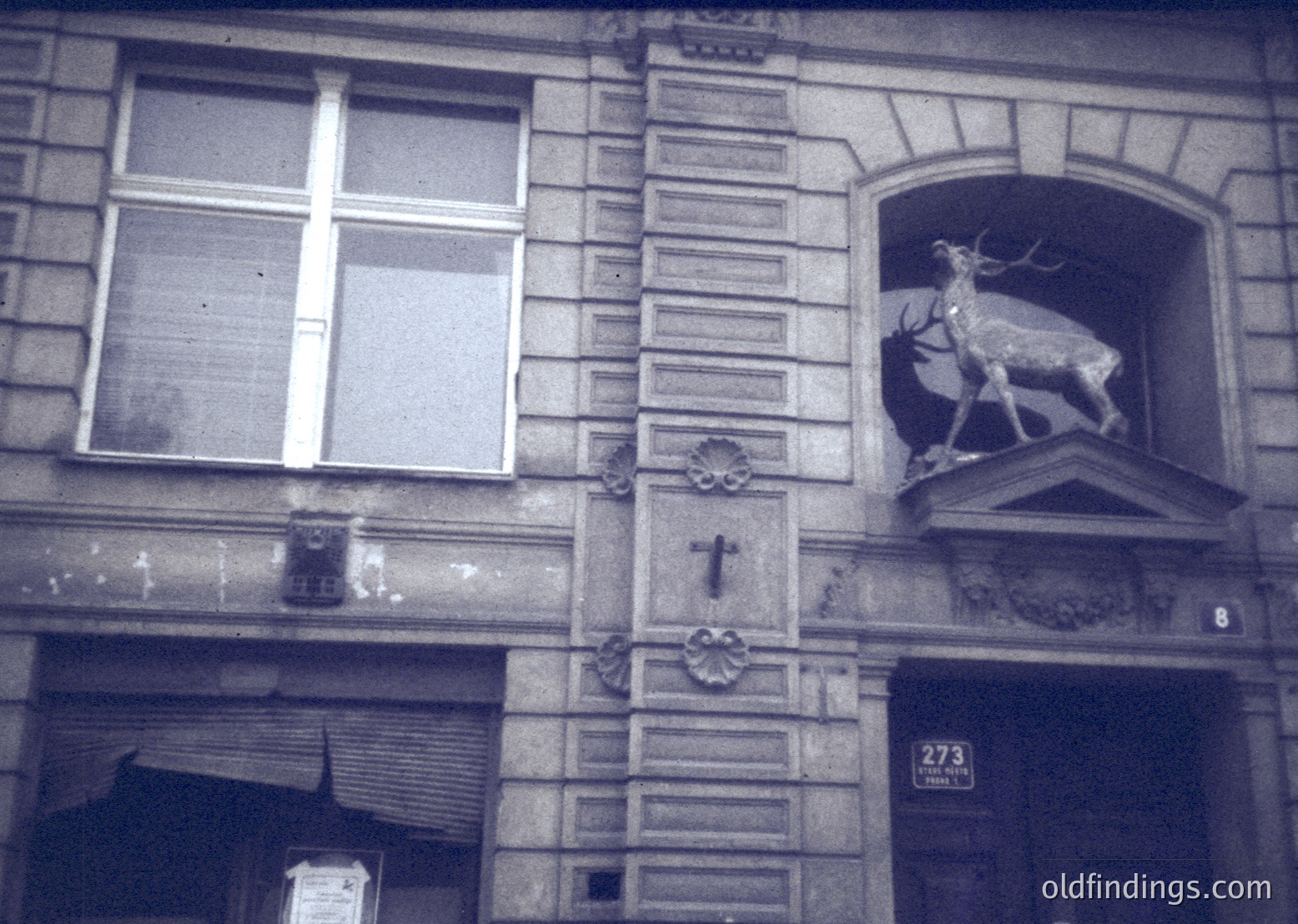 Black-and-white facade of a European building (likely 19th–early 20th century) featuring ornate stonework: a relief deer sculpture above a wrought-iron cross, decorative floral motifs, and a dated plaque (1877). Architectural style suggests historic urban setting.
