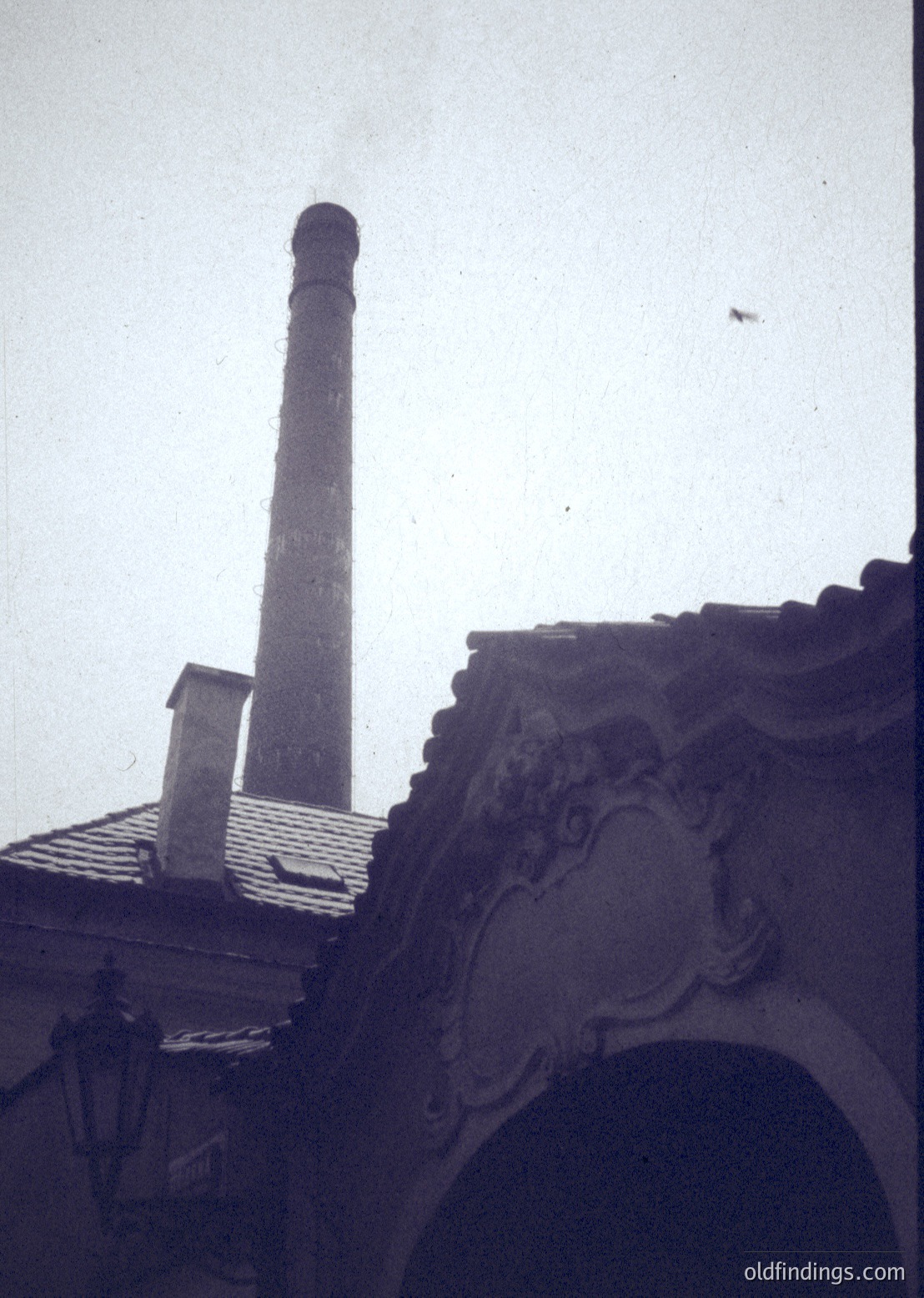 Black-and-white industrial photograph featuring a tall brick smokestack against a hazy sky, with ornate architectural details (scrollwork) on a rooftop edge in foreground. Likely 1940s–1960s European factory setting.