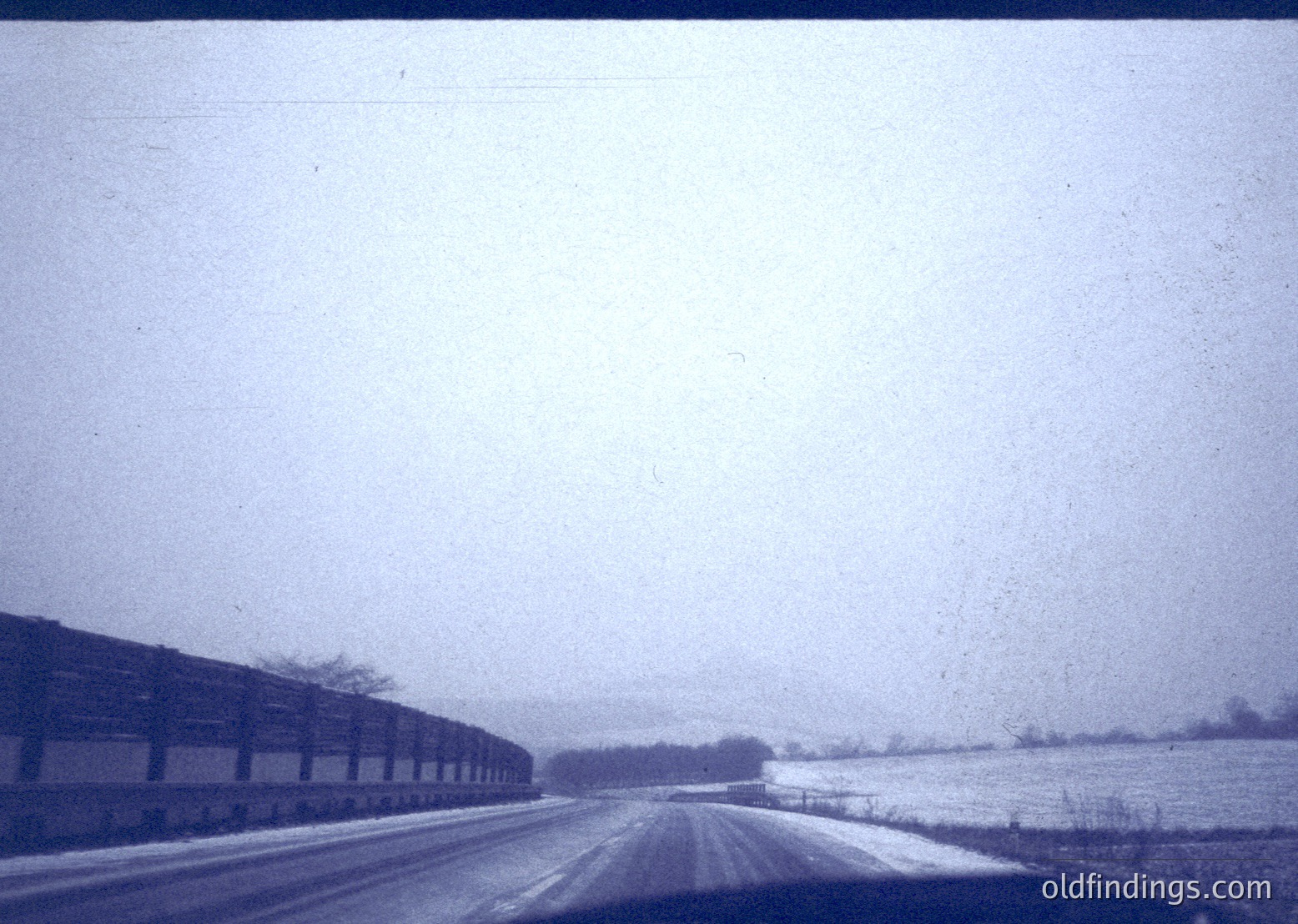 Vintage black-and-white photo of a rural road flanked by snow-covered fields and leafless trees. Industrial brick buildings line the left side, suggesting agricultural or industrial use. Overcast sky enhances the wintry atmosphere. Likely late 20th century () in a temperate climate region.