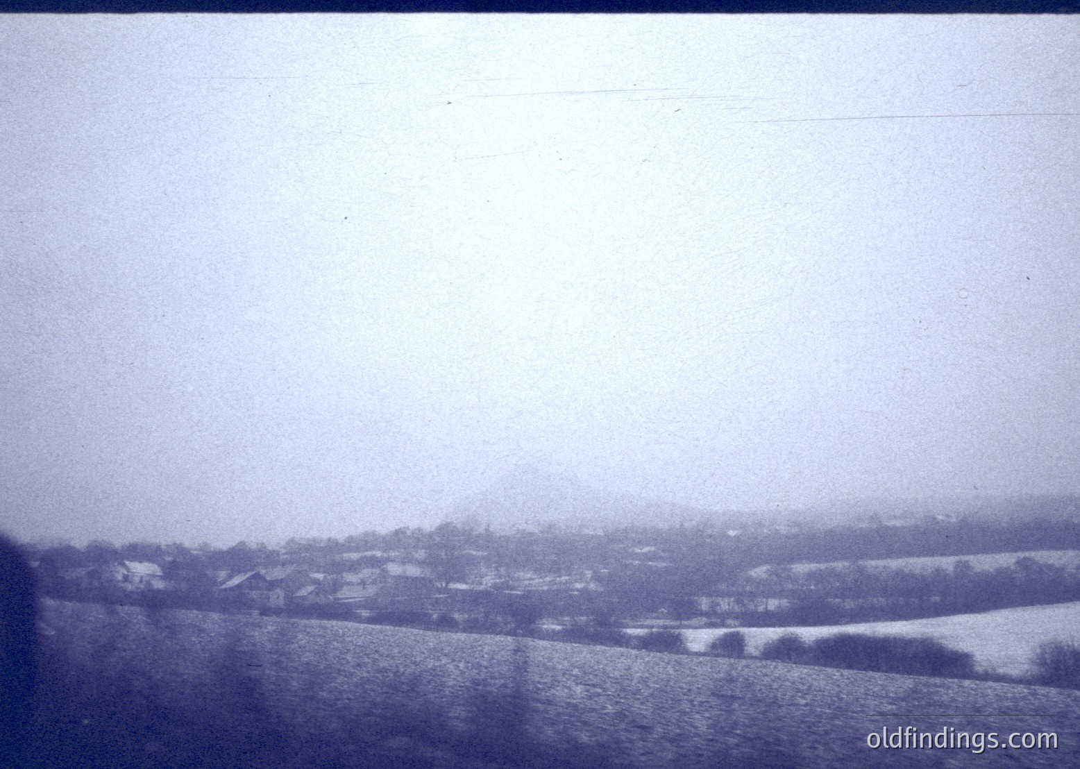 Vintage black-and-white landscape featuring snow-covered fields and distant village. Low-lying hills and misty horizon suggest early 20th-century rural Europe. Faded film grain enhances nostalgic vintage aesthetic.