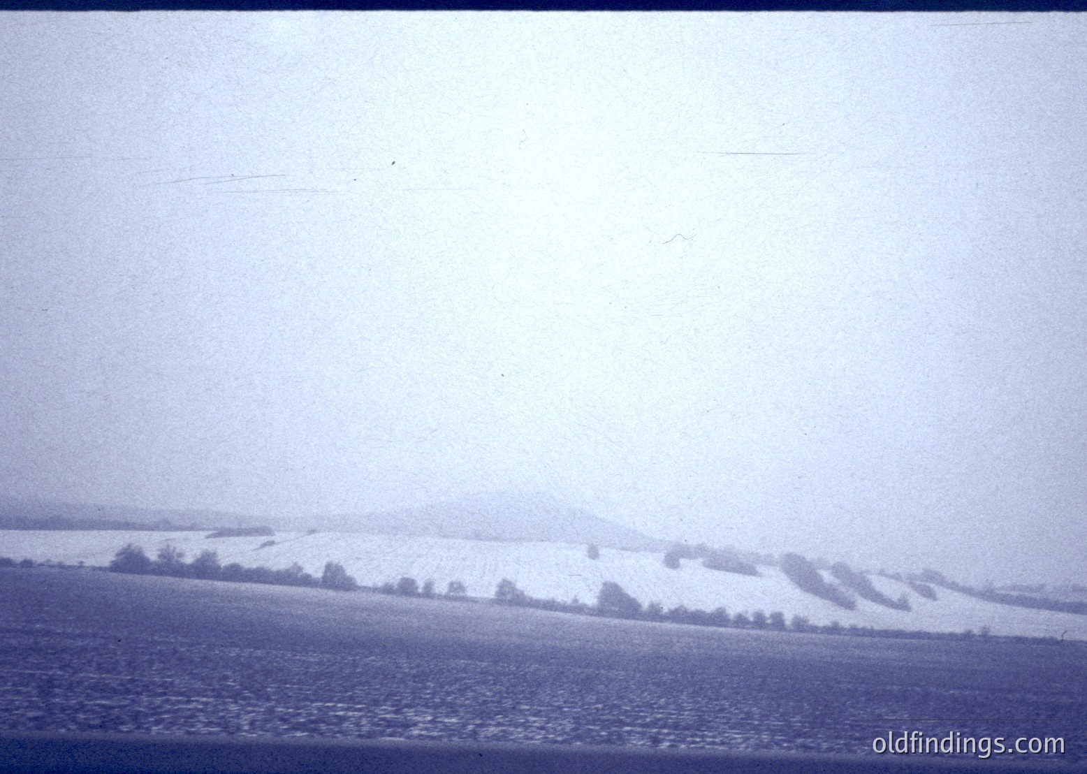 Vintage black-and-white landscape showing snow-covered hills and sparse tree line under overcast skies. Flat, open field dominates foreground, suggesting rural agricultural land. Likely mid-20th century due to grainy film texture.