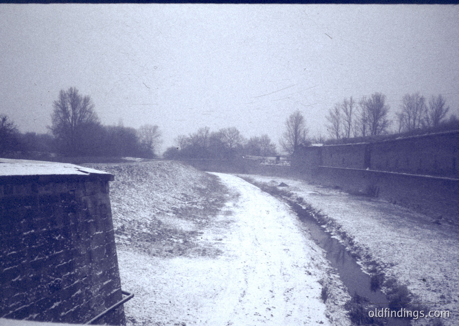 Mid-20th century black-and-white rural roadside scene with light snow. Concrete guardrail and utility pole flank a narrow, snow-covered gravel road. Bare trees and distant farm buildings suggest agricultural setting. Foggy, overcast skies enhance nostalgic atmosphere.