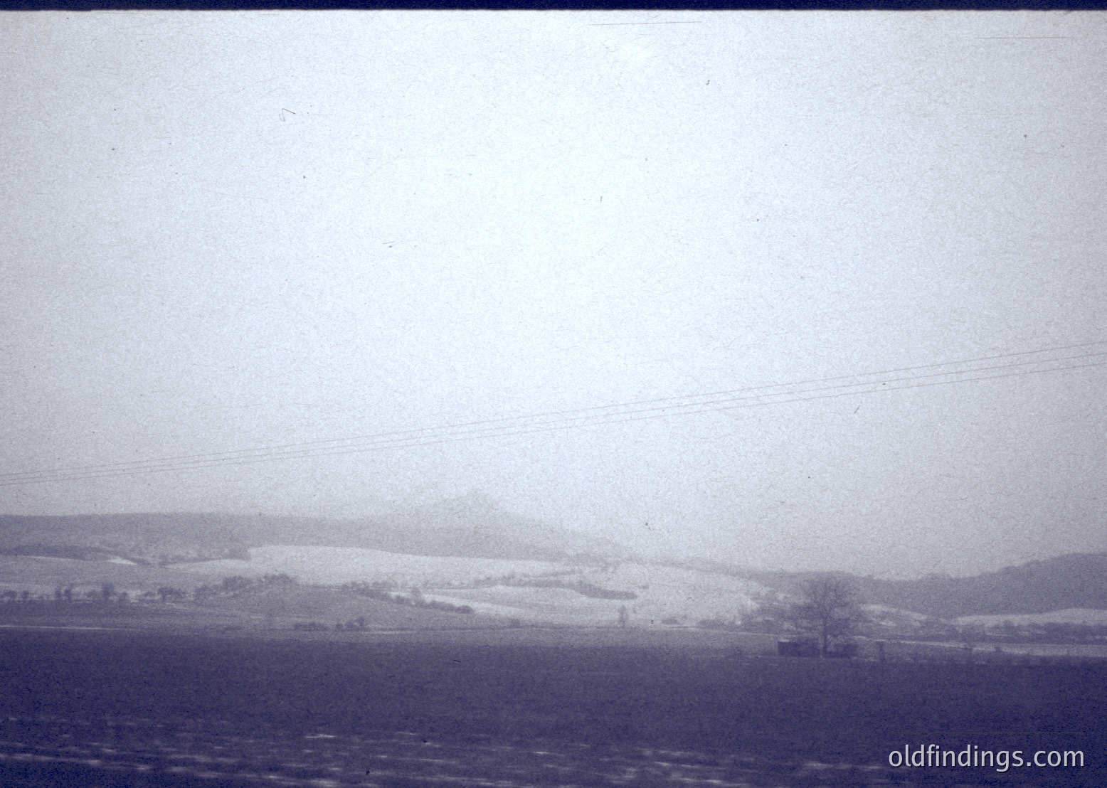 Vintage black-and-white landscape showing snow-covered hills under overcast skies. Distant farmland and sparse trees suggest rural setting. Low-contrast, grainy texture indicates mid-20th century photography.