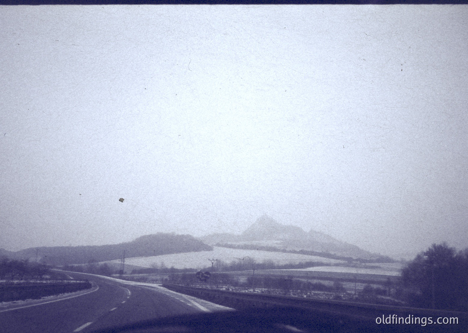 Vintage black-and-white roadside scene with misty mountains in background, likely mid-20th century. Curved road flanked by sparse trees and open fields, hinting at rural isolation. Fog obscures distant peaks, creating atmospheric depth.