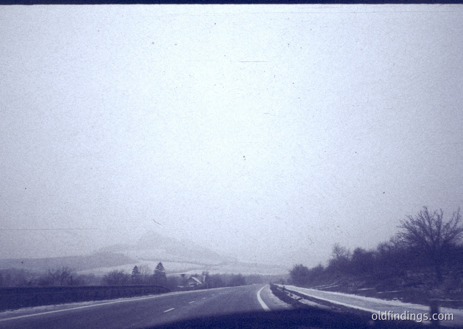Vintage black-and-white roadside scene showing a two-lane highway with faint snow remnants. Distant, mist-covered mountain range and sparse winter trees frame the horizon. Likely mid-20th century, rural setting.