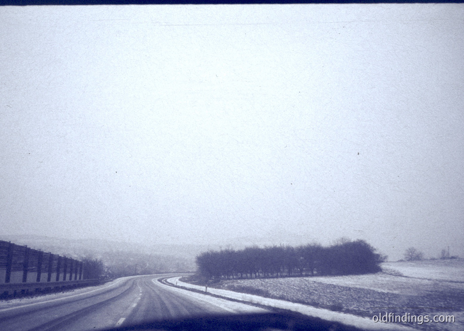 Blurred vintage roadside view with light snowfall, featuring a single-lane road bordered by industrial buildings on left and open fields on right. Monochrome, grainy texture suggests mid-20th century photography.