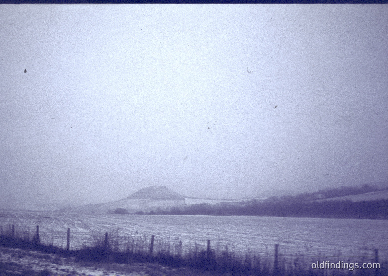 Vintage black-and-white landscape showing mist-covered rolling hills and open fields. Faint fence lines and sparse vegetation suggest rural agricultural land. Fog obscures distant horizon, creating atmospheric depth. Likely early-to-mid 20th century due to photographic style.