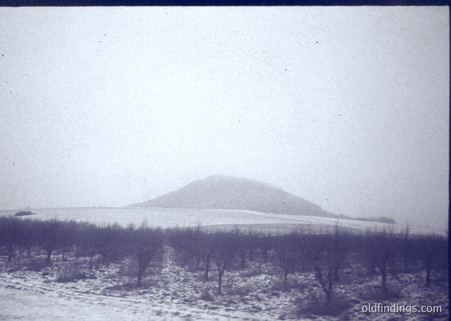 Vintage sepia-toned landscape featuring a snow-covered hillside with sparse, leafless trees in foreground. Overcast sky obscures distant horizon, creating muted tones. Likely early-to-mid 20th century rural setting.