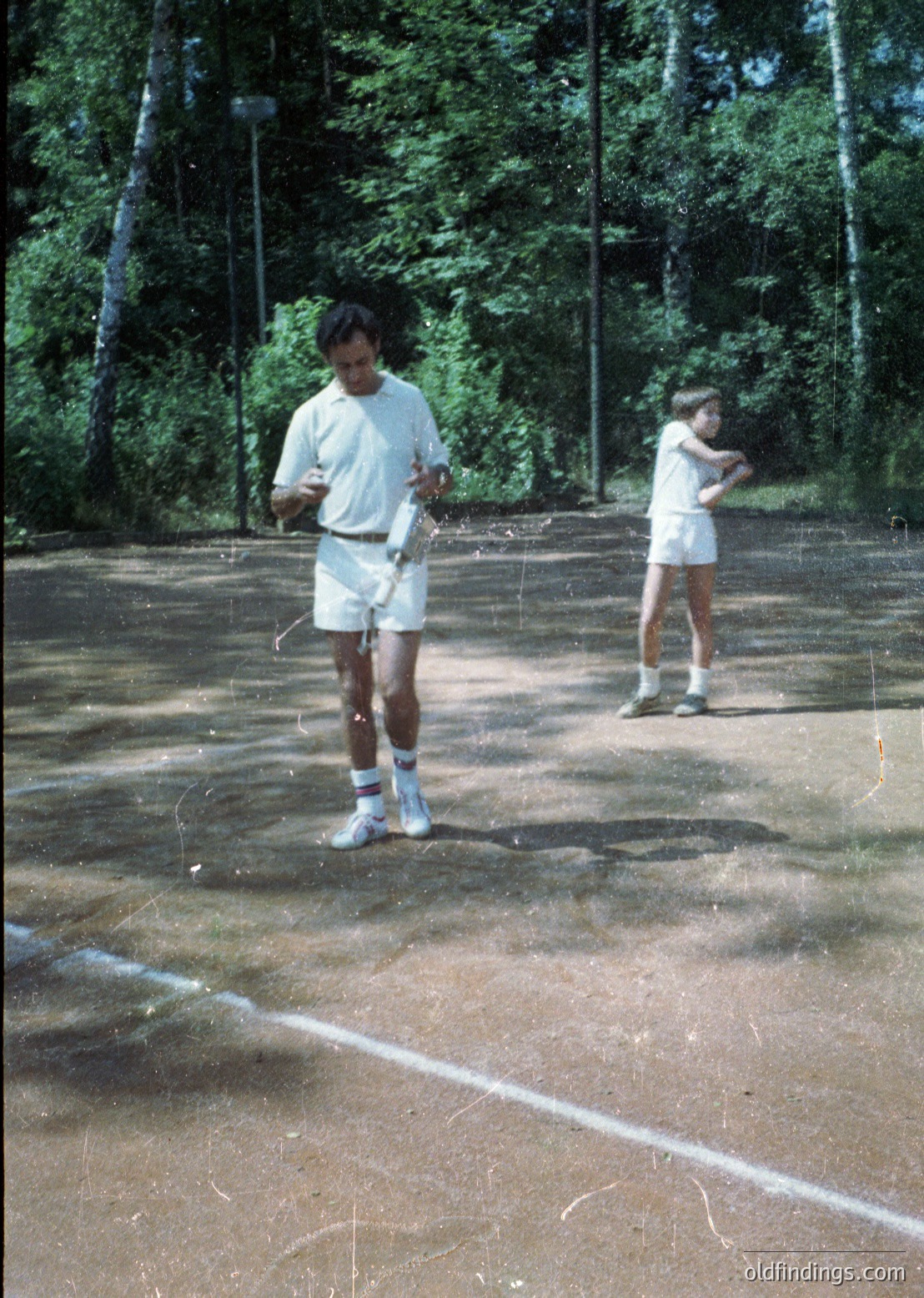 Two individuals in 1970s-style athletic wear—white shirts, shorts, and sneakers—play tennis on a worn outdoor court surrounded by dense forest. The court’s faded lines and rough surface suggest frequent use. Likely mid-20th century, possibly North America or Europe.