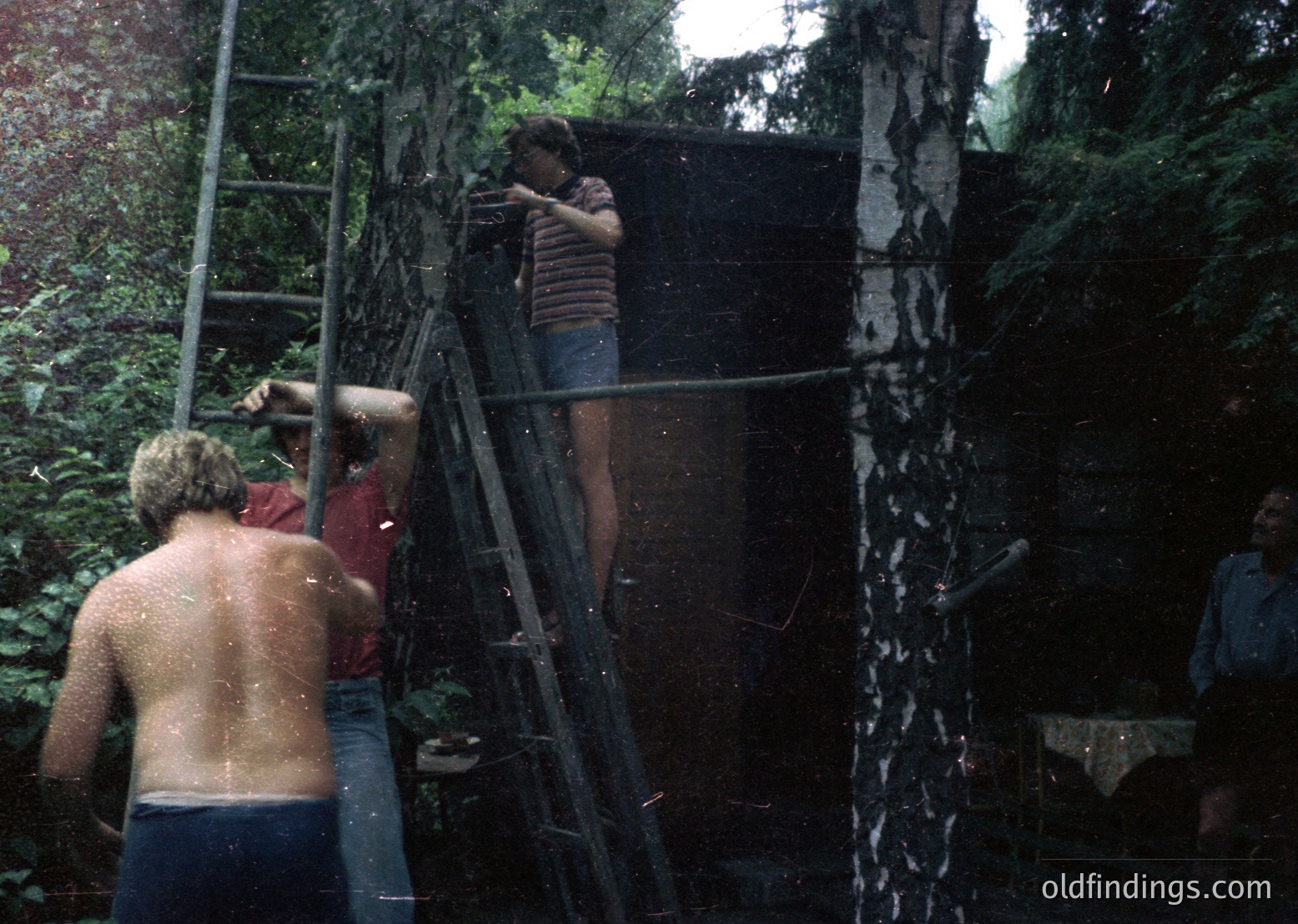 Vintage 1970s-style group water fight under a cascading waterfall in a lush forest. Three shirtless men and one partially visible person engage playfully, using a ladder and buckets. Casual summer attire—bell-bottoms, striped shirts—evokes retro beach/outdoor party vibes. Ideal for nostalgia, stock themes of youth culture, or historical research on 20th-century leisure.