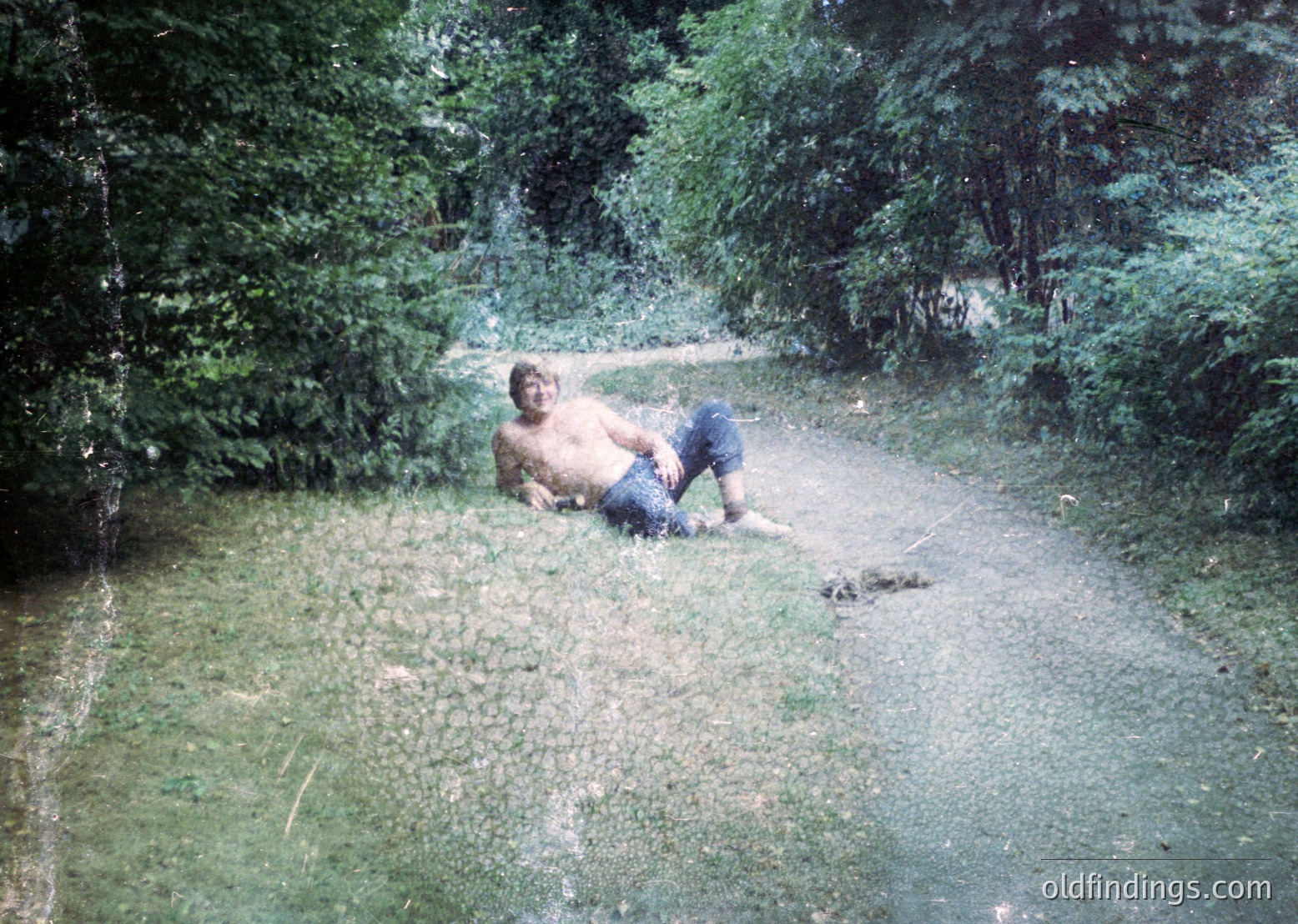 Vintage photo of a shirtless man sitting on wet grass beside a shallow stream in a forested area, mid-20th century. Lush greenery and a dirt path frame the scene, suggesting a rural or park setting.