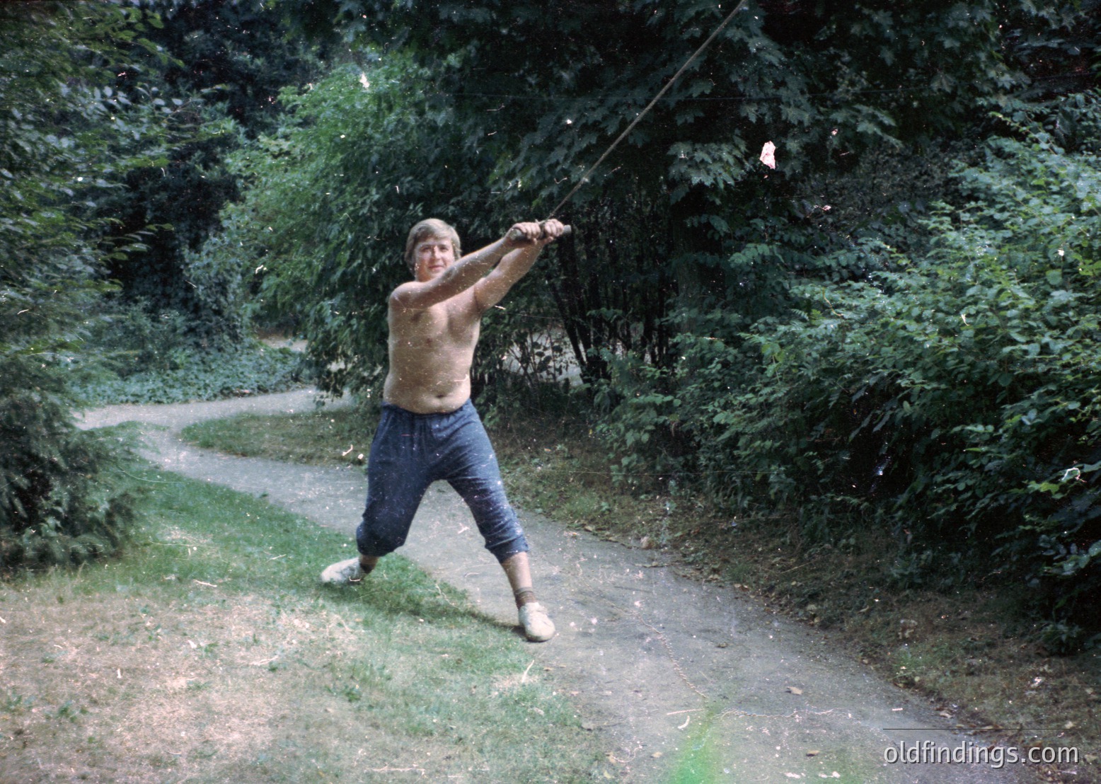Shirtless man mid-action swinging a long wooden pole in a rural, wooded path, 1970s. Casual attire includes rolled-up trousers and worn sneakers. Lush greenery and dirt road suggest outdoor recreation or folk activity.