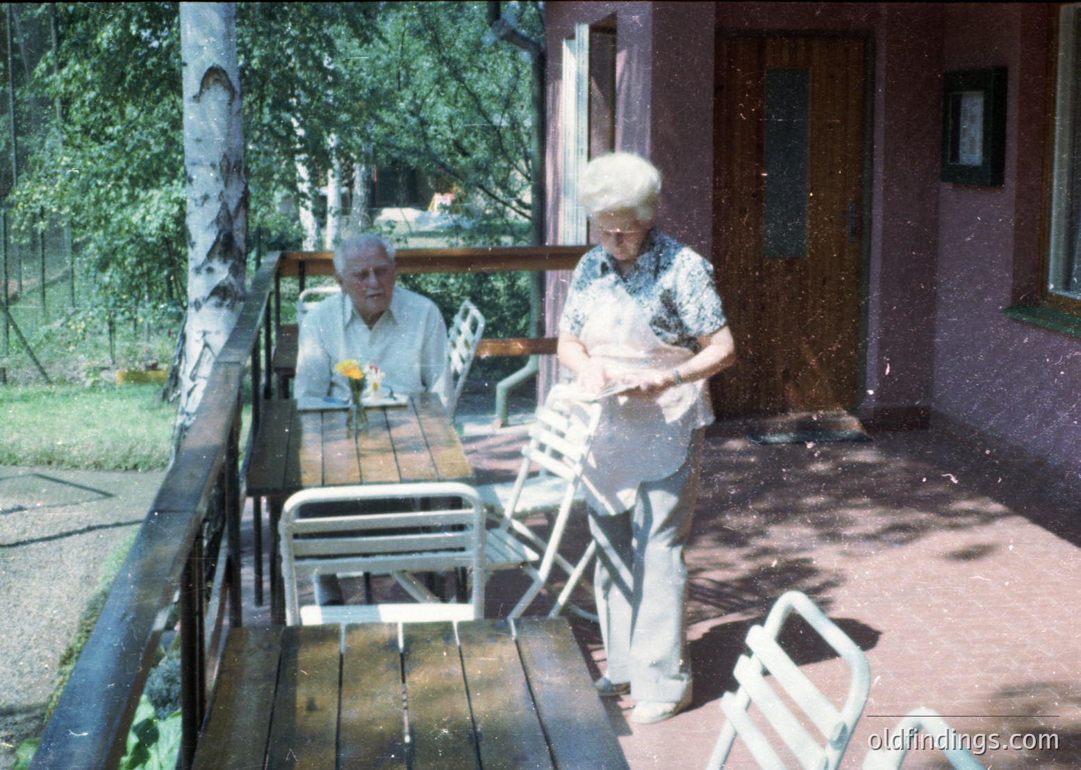 Vintage outdoor scene featuring two elderly individuals on a wooden balcony patio. Man seated at a table with a drink, woman standing beside a chair holding a book. Mid-century metal furniture and lush greenery in background. Likely Eastern European resort setting, 1960s-1970s.